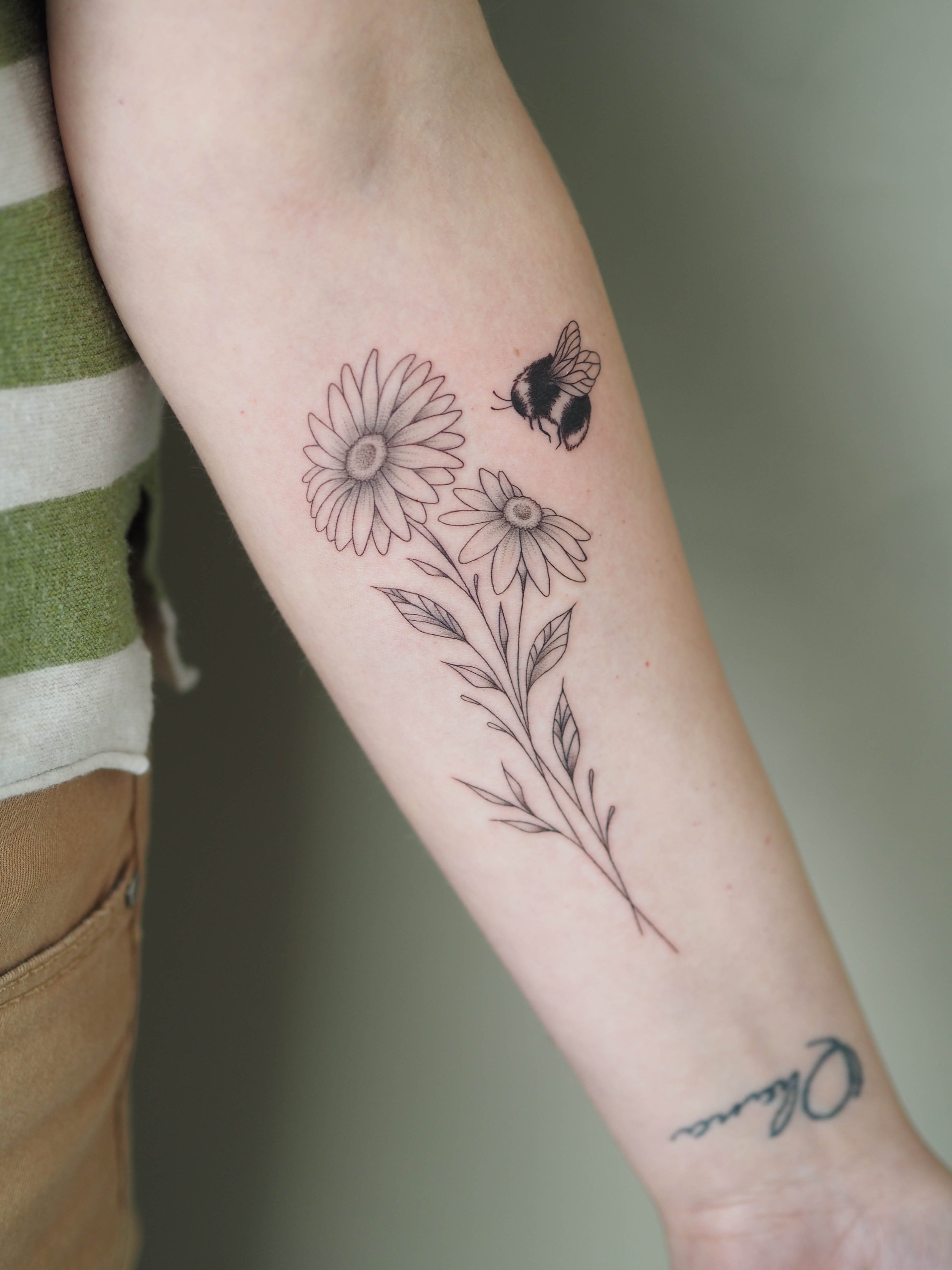 Tattoo eines Blumenstraußes mit zwei Gänseblümchen und einer Biene auf dem Unterarm.