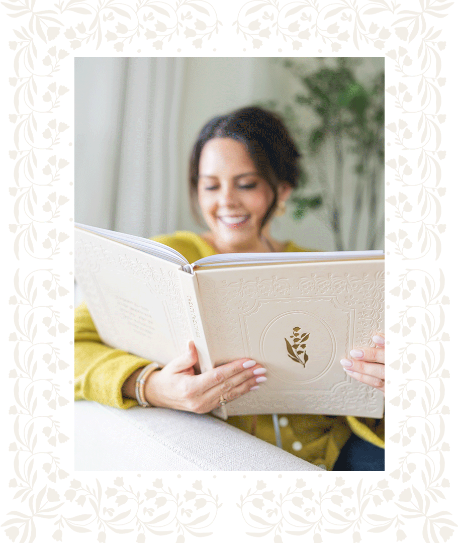 A woman with dark hair, wearing a yellow top, is sitting on a light-colored sofa, reading a large book with a cream-colored cover that has a decorative border and a gold leaf emblem. She is smiling and appears happy, with a blurred background featuring a plant.