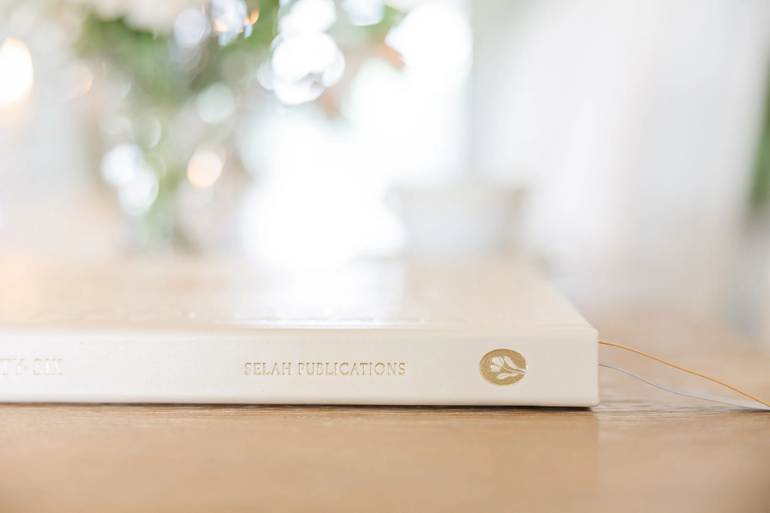 Close-up of a white book titled 'Selah Publications' resting on a wooden surface, with a gold logo of a leaf on the spine. Blurred background with Christmas lights and greenery.