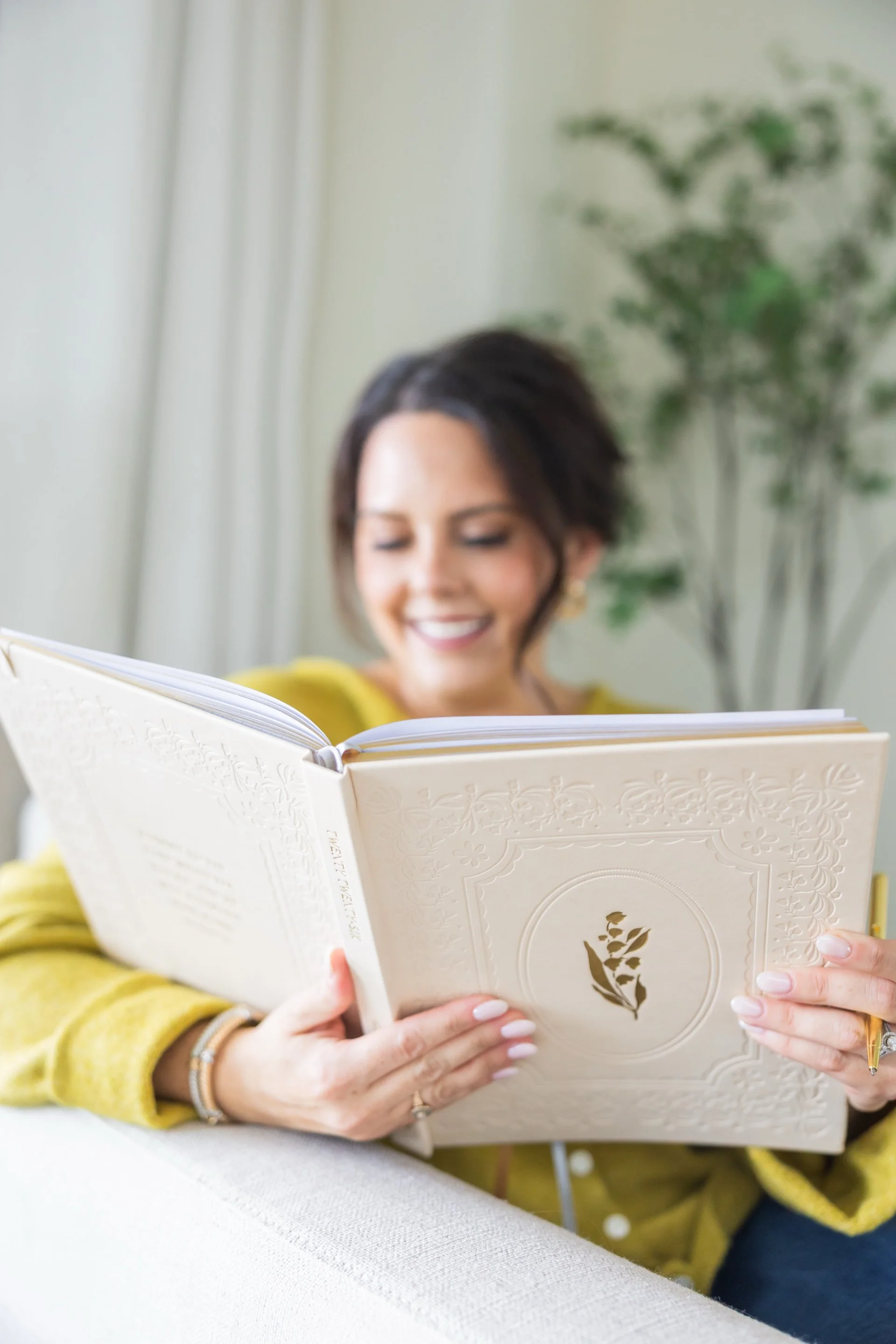 Woman smiling while reading a book with a floral design on the cover, sitting on a white couch with a green plant in the background.