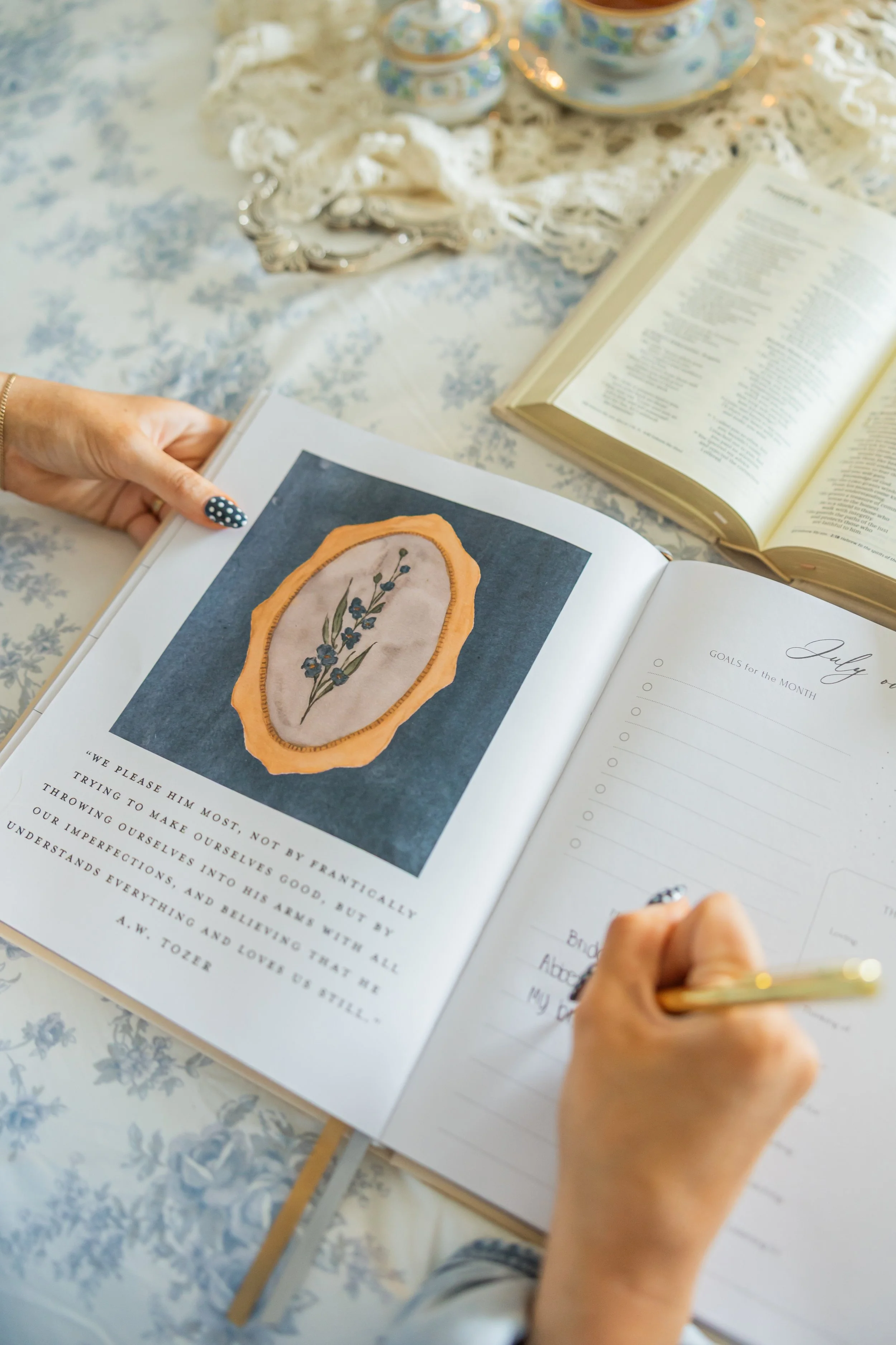 Person writing in a planner with a floral design, surrounded by tea set, open books, and vintage jewelry on a lace-covered table.