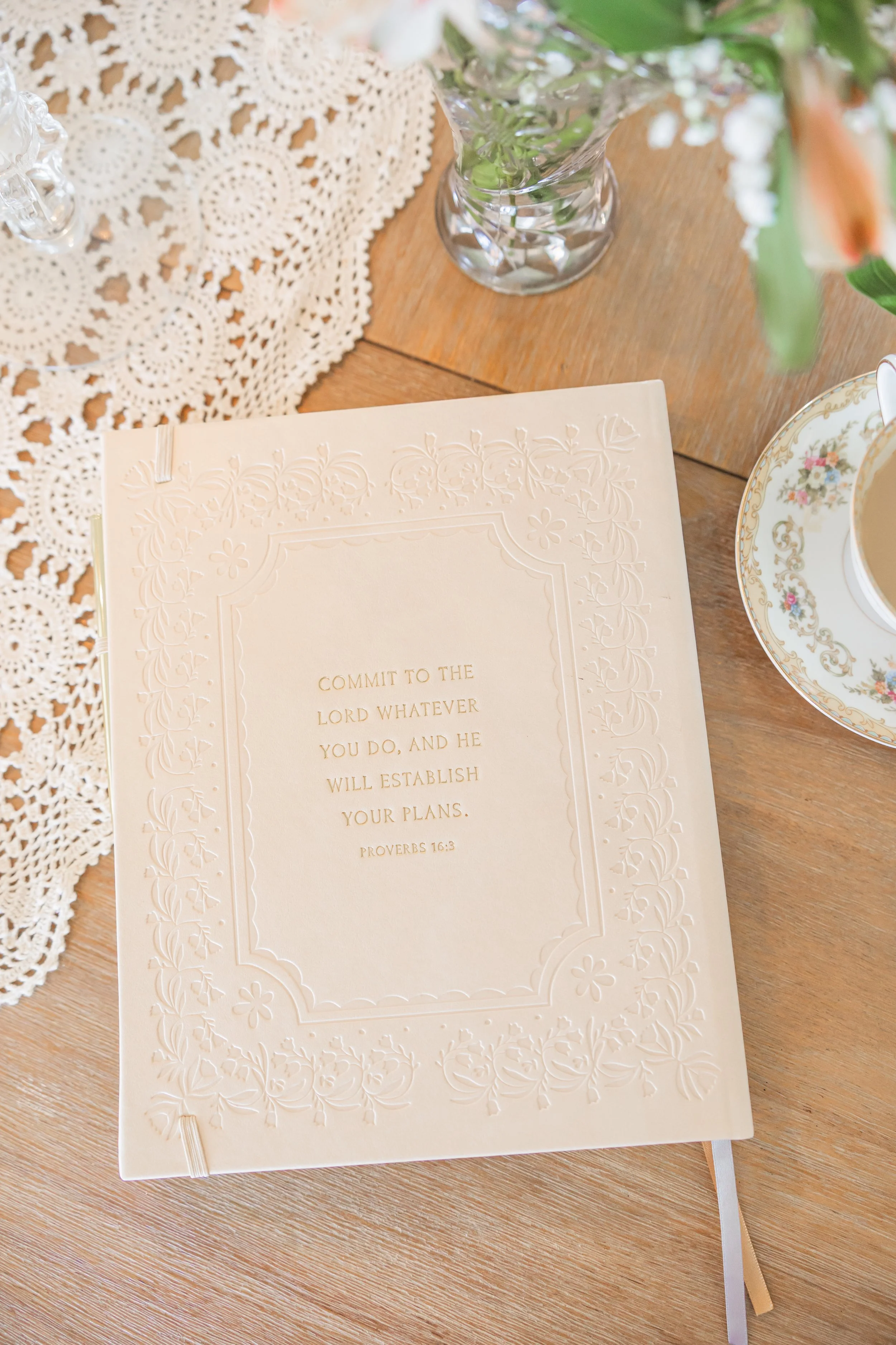 A prayer journal with embossed floral border and gold text on a wooden table, surrounded by a doily, a vase with pink and white flowers, and a floral teacup with tea.