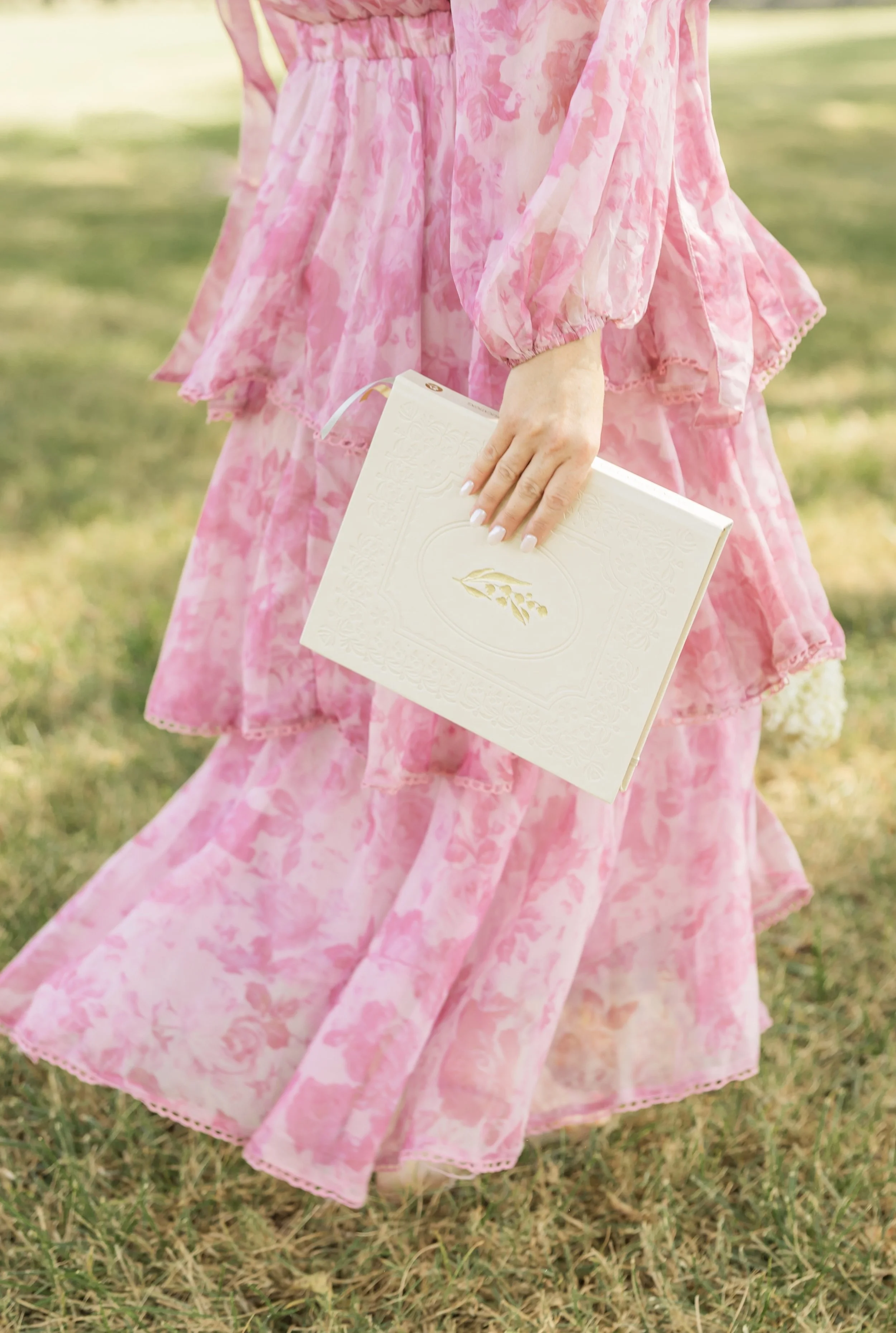 A woman holding a white book or journal with embossed details, wearing a flowing pink floral dress, standing on green grass outdoors.