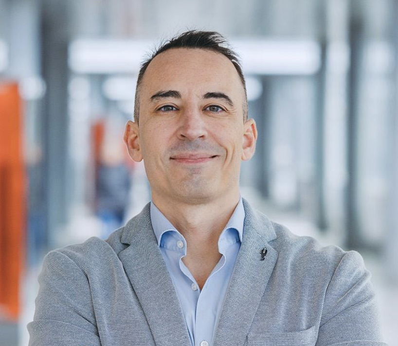 Hombre con cabello oscuro y peinado hacia un lado, vestido con un saco gris y una camisa azul claro, sonriendo en un ambiente de oficina moderno y bien iluminado.
