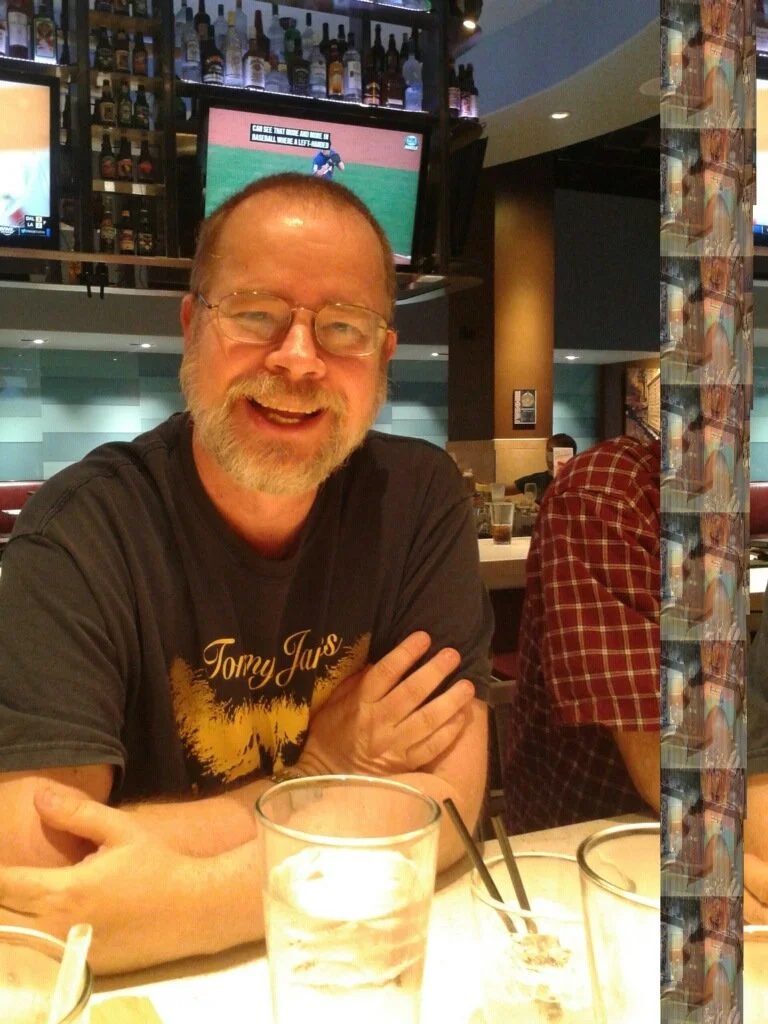 A middle aged man with a grey beard, short cropped hair and glasses can be seen smiling widely at the camera. He appears to be seated in a bar or restaurant. His arms are crossed, resting on a table, a wall of bottles & TVs behind.
