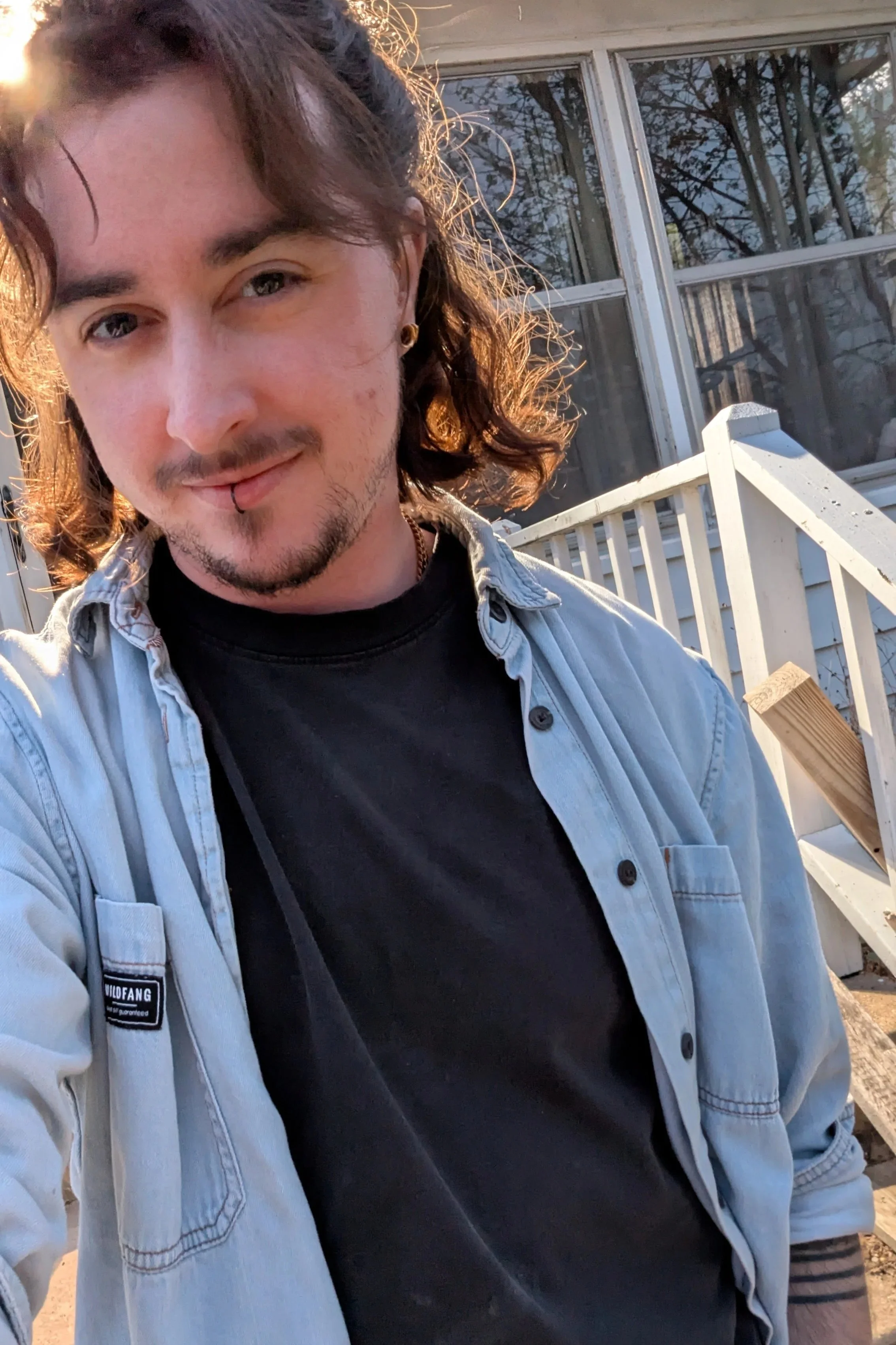 cora has shoulder-length curly reddish brown hair, short facial hair, and dark eyes. They are wearing a blue denim shirt over a black tshirt. The sun is behind them and a white house is visible over their shoulder.
