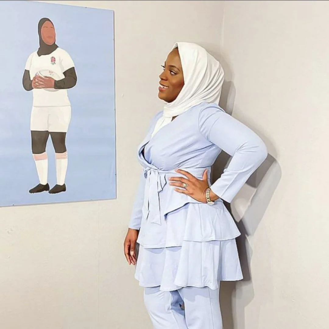 A woman wearing a light blue dress with a white headscarf, standing sideways with one hand on her hip, next to a painting of a rugby player in a white uniform holding a rugby ball.