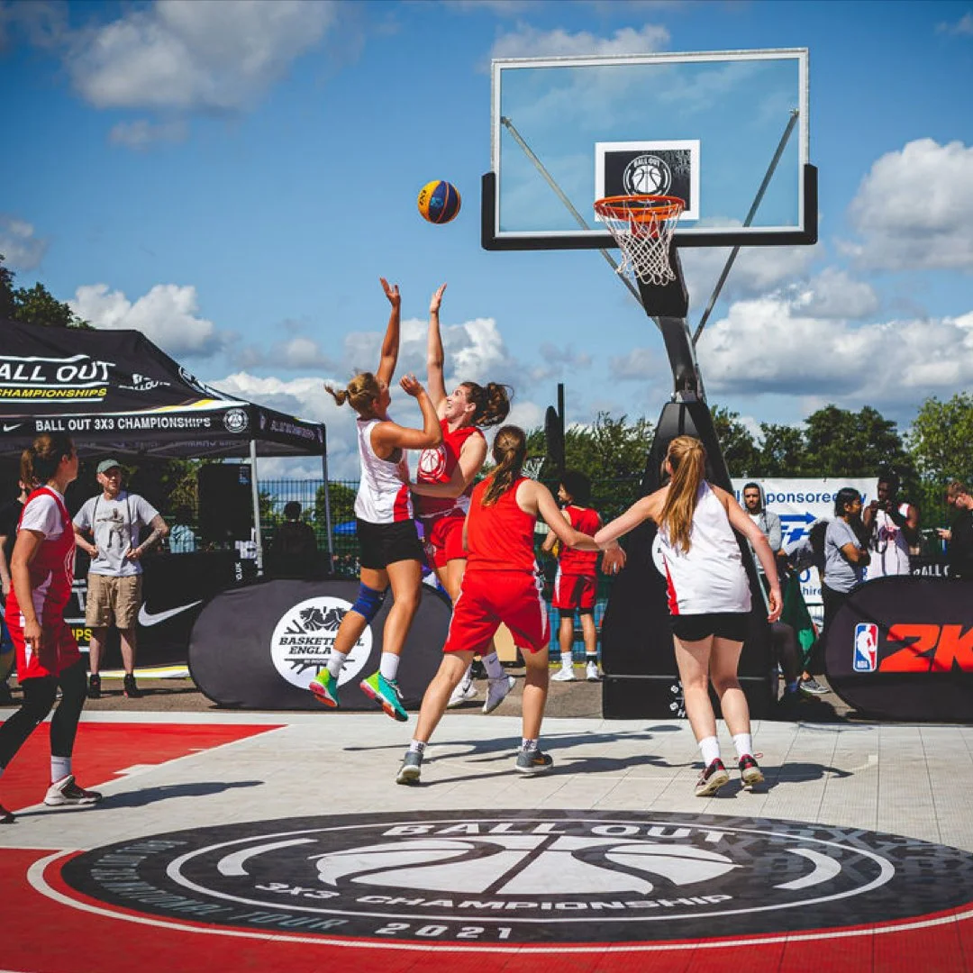 Women playing basketball on an outdoor court during a tournament, with some players jumping for the ball near the hoop. The court has a logo that reads 'Ball Out 3x3 Championship 2021' and is set up with banners and a tent in the background.