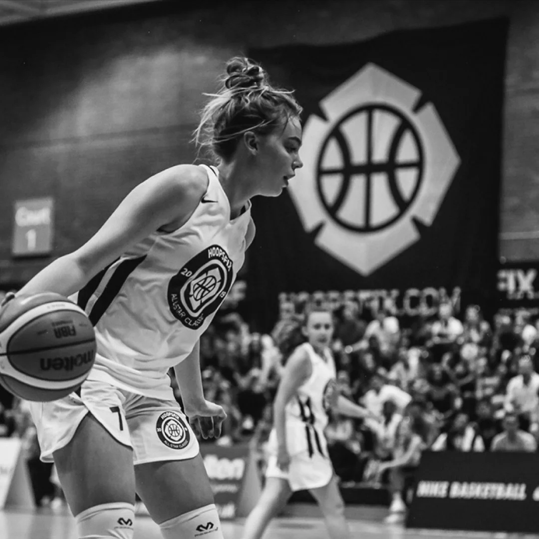 A female basketball player dribbling in a game, wearing a jersey with the logo 'Hoopity' and surrounded by spectators.
