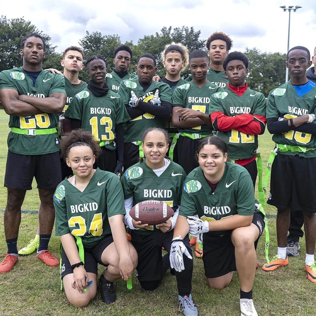 A youth football team in green jerseys with 'BIGKID' printed on them, posing outdoors on a football field. The team includes both boys and girls, with some kneeling and others standing, holding a football and wearing bright athletic shoes.