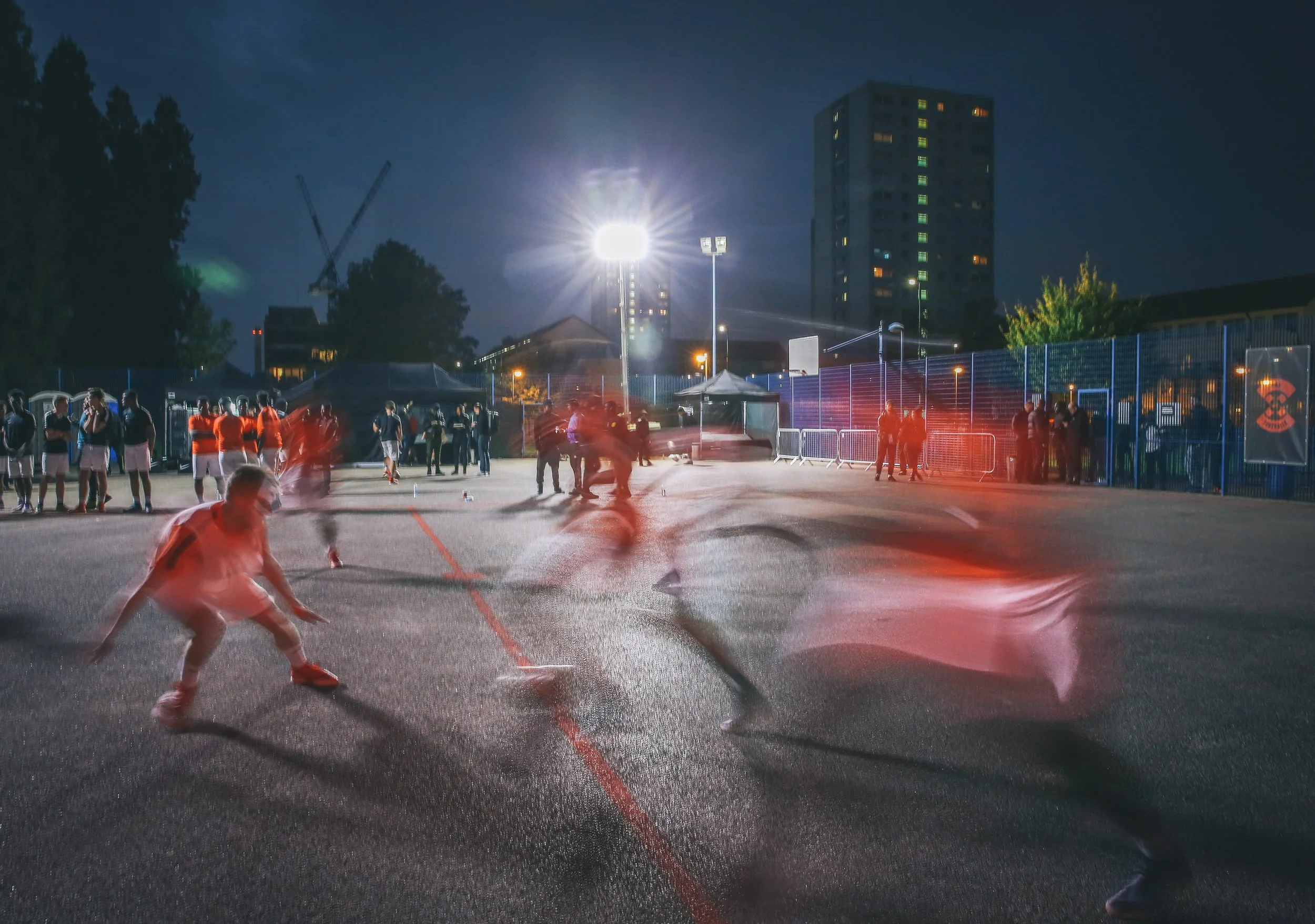 Nighttime scene on a sports court with children and adults playing and watching, illuminated by floodlights, with onlookers and tall buildings in the background.