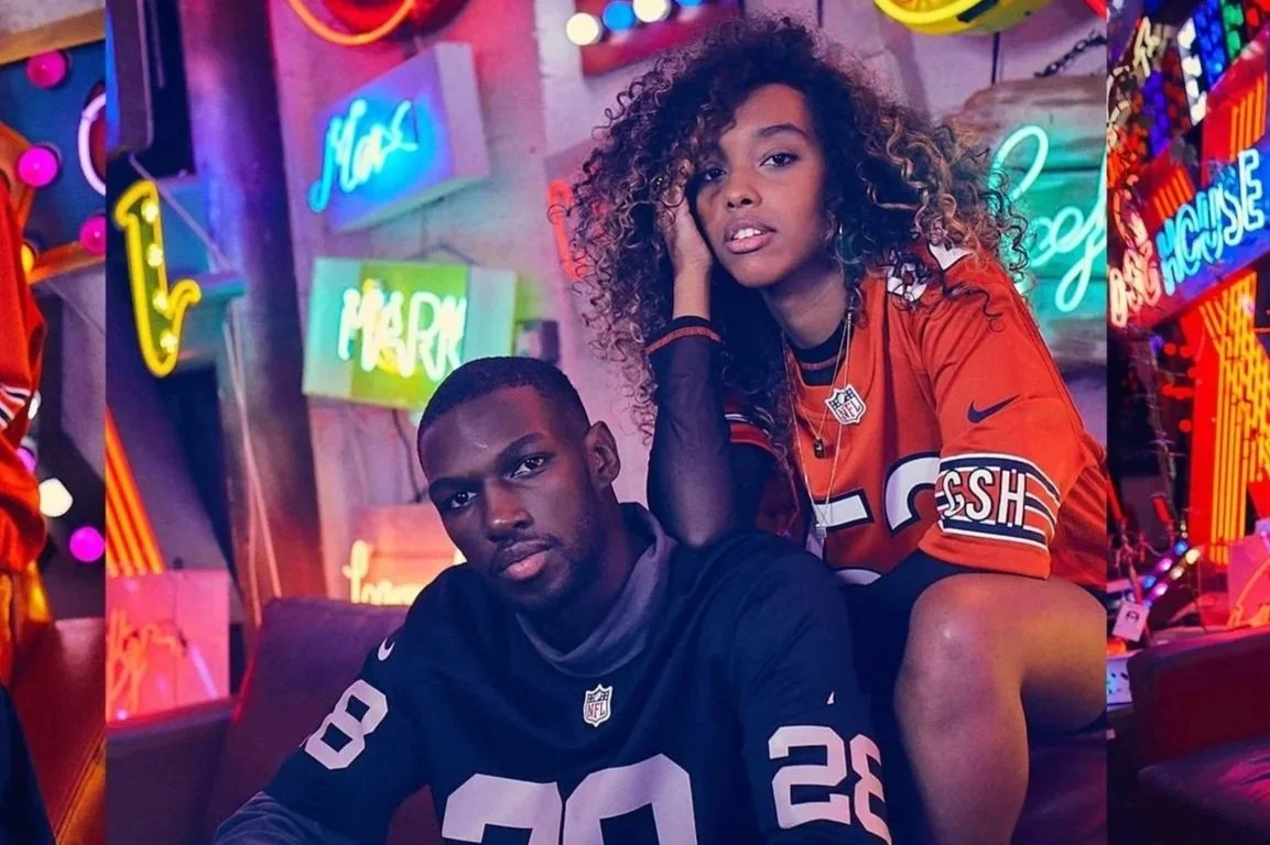 Two young adults, a man and a woman, are posing in front of neon and colorful signs in an indoor setting. The man is sitting on a couch wearing a dark NFL jersey, and the woman is sitting on his knee, wearing an orange NFL jersey, with curly hair, touching her face.