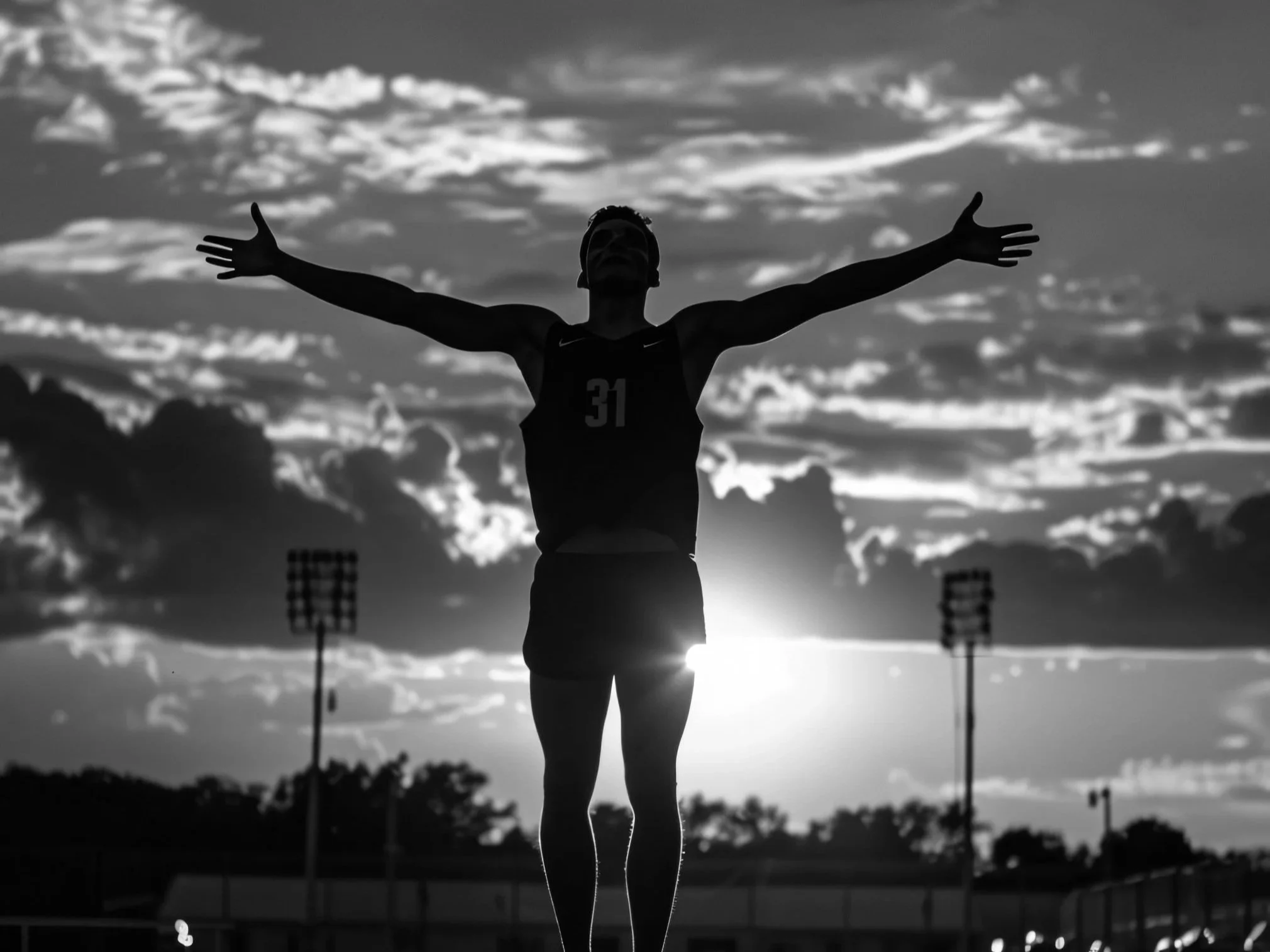 Silhouette of a person with arms outstretched standing outdoors at sunset or sunrise, with stadium lights in the background.