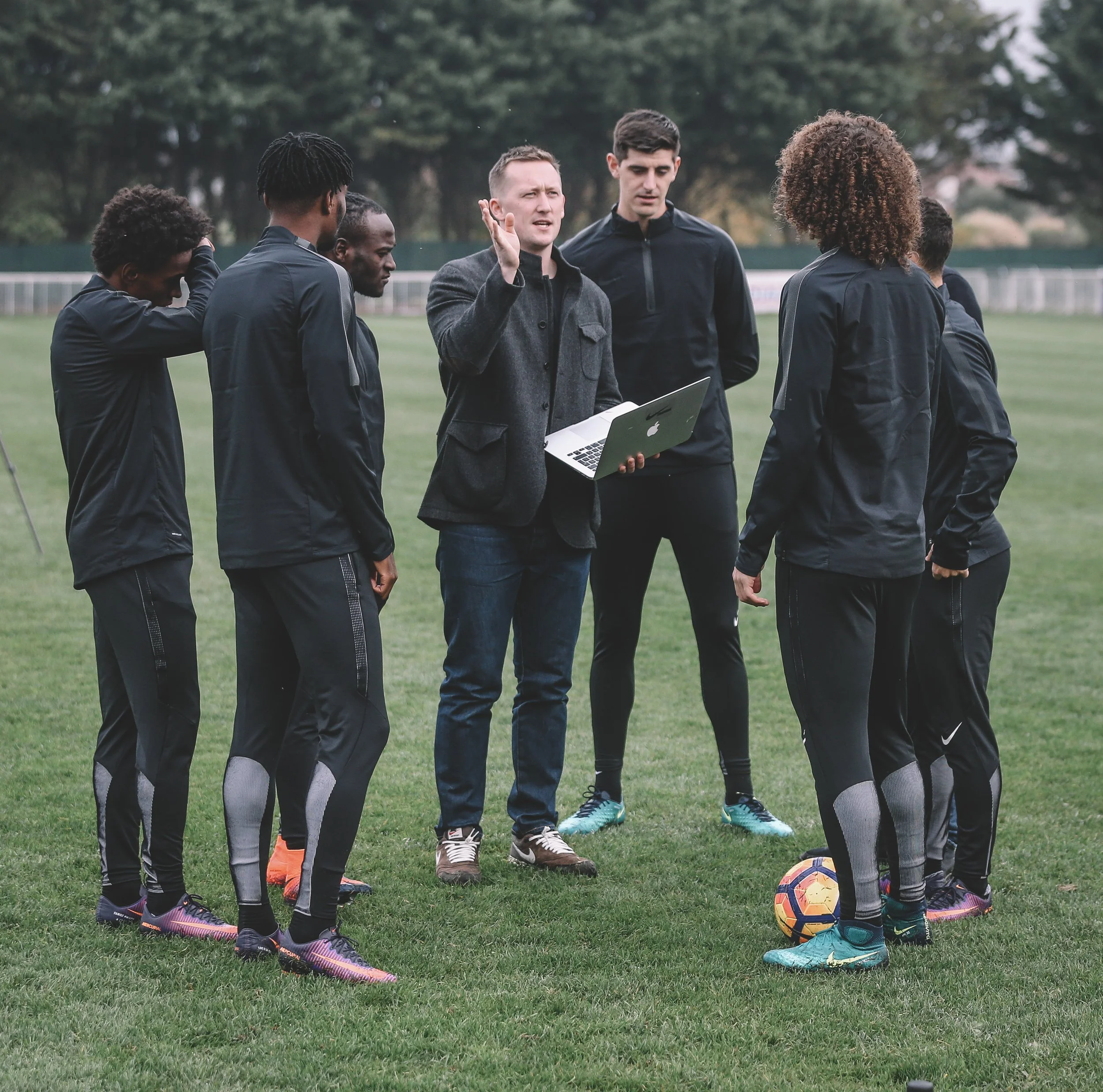 Richard Frost giving instructions to a group of players on a soccer field, with a laptop in hand and a soccer ball at their feet.