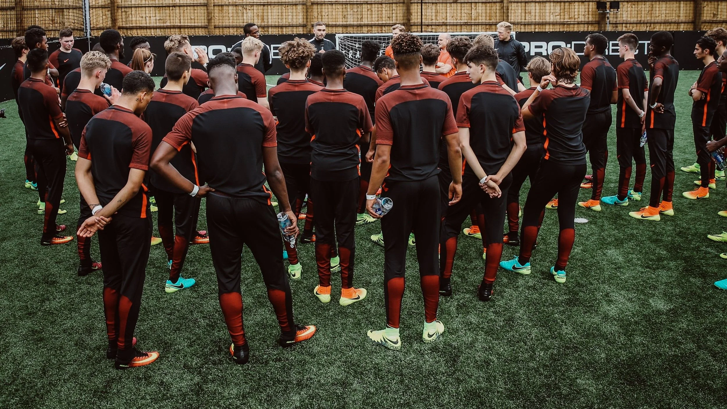 A group of young male soccer players in black and red uniforms gathered on a training field, listening to their coach during a team meeting.