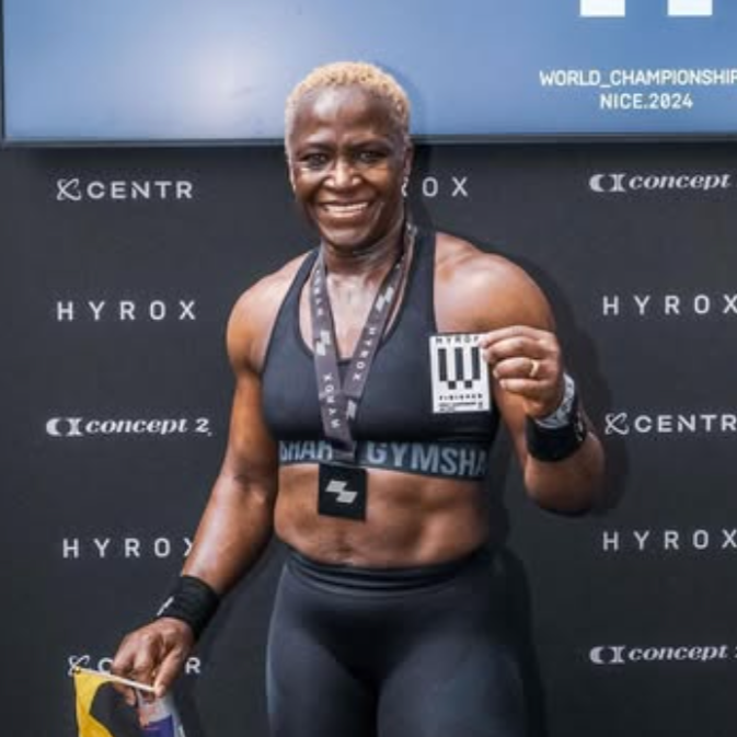 A female athlete smiling and holding a race bib, standing in front of a sponsored backdrop at the 2024 World Championships in Nice, wearing a sports bra and leggings with a medal around her neck.