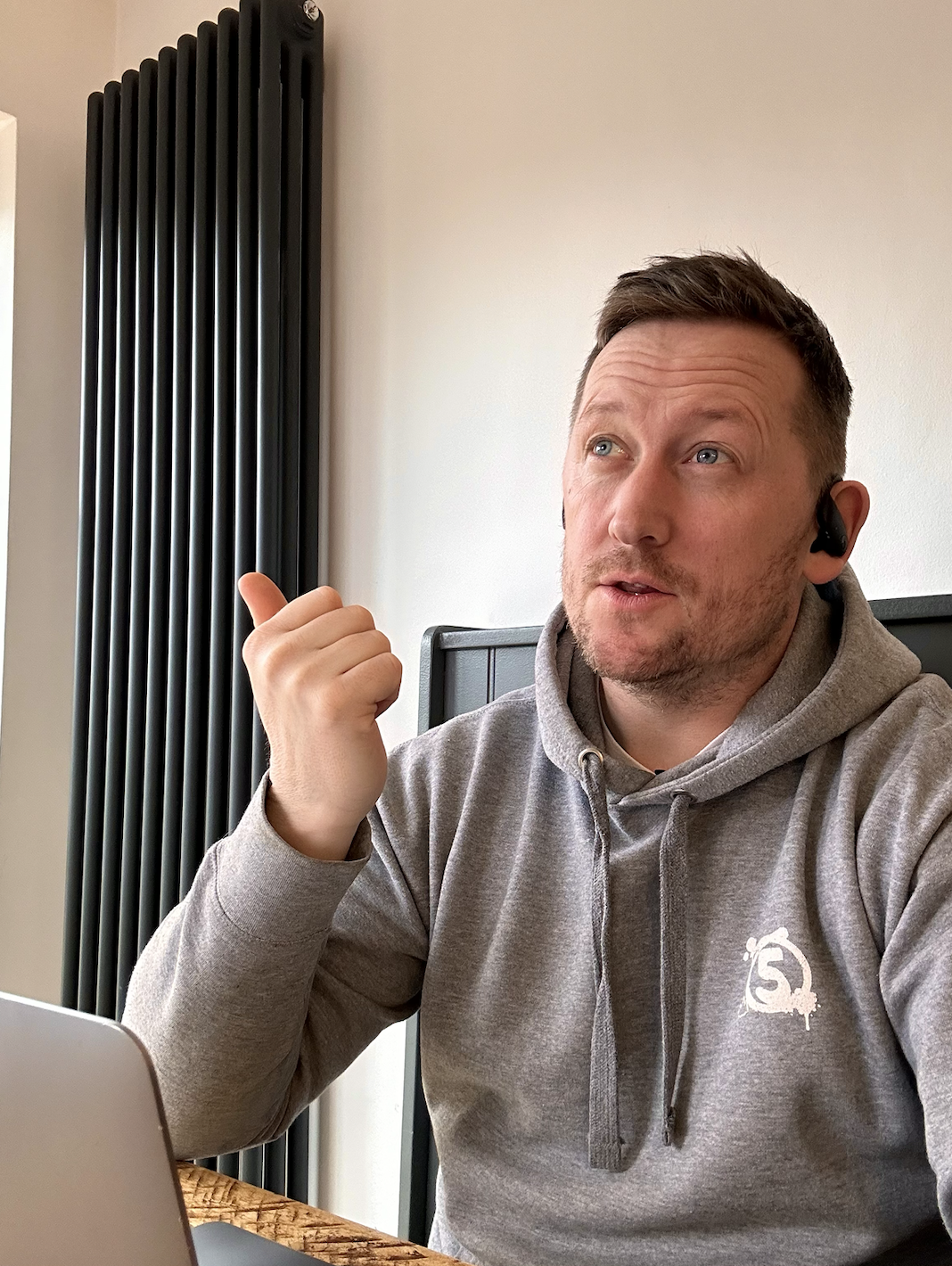 Richard Frost wearing a grey hoodie with a logo, sitting at a table with a laptop, gesturing with his hand, and wearing a Bluetooth earpiece, in a room with a radiator and plain wall.