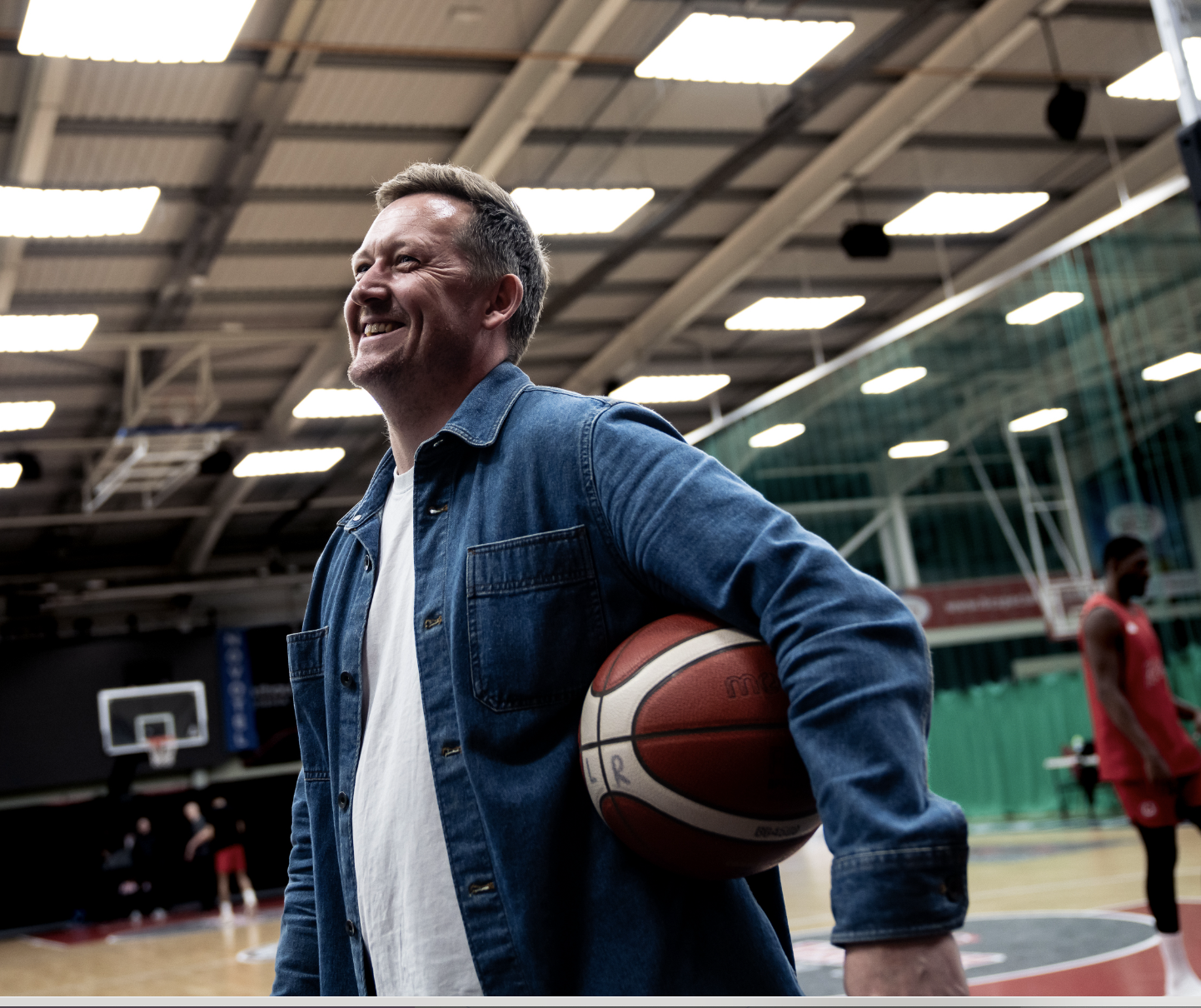 Richard Frost holding a basketball in a gymnasium with basketball players and hoops in the background.
