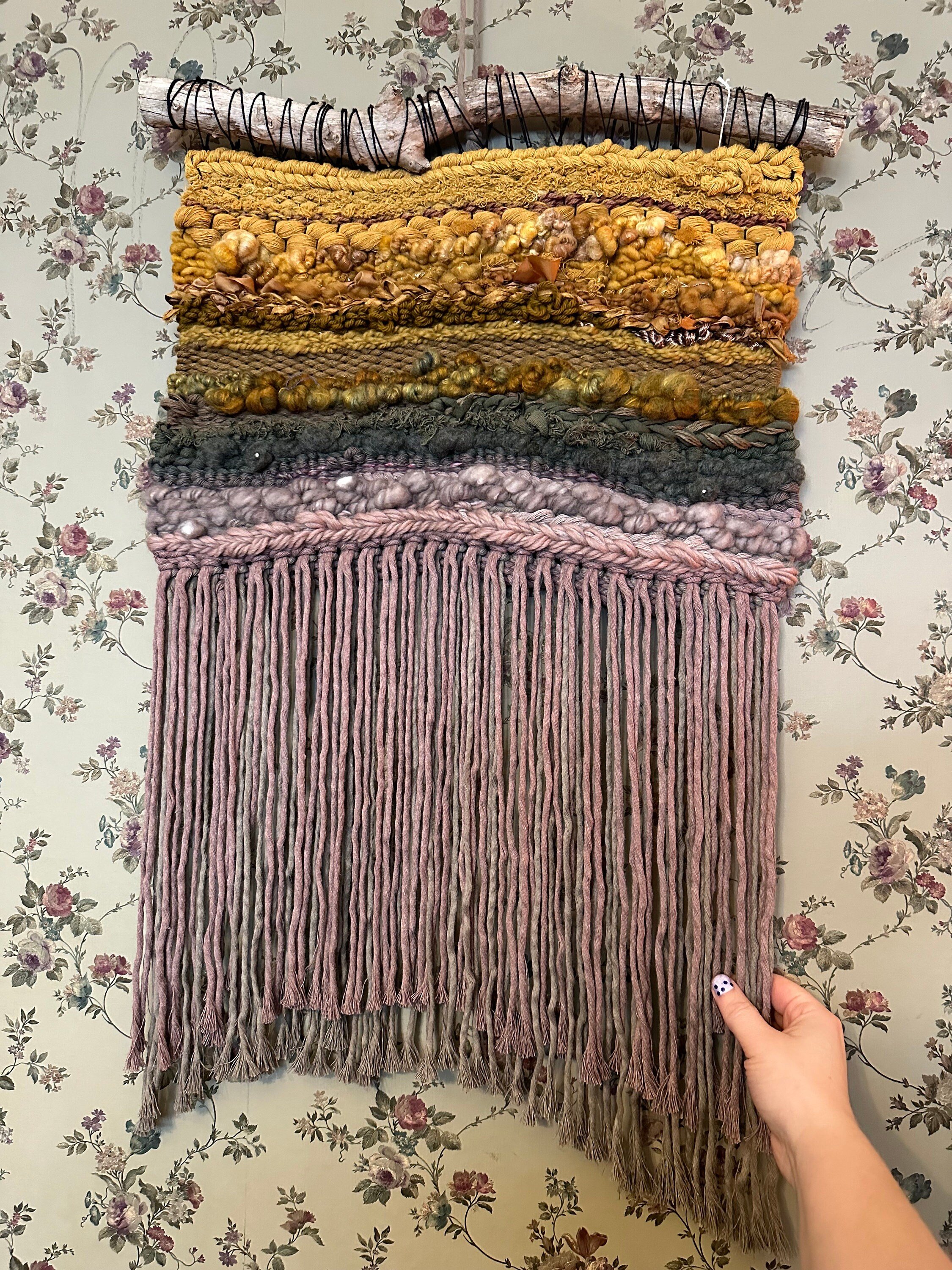 handmade woven tapestry in golds, greens, taupes and mauves hangs on a driftwood; white hand is holding fringe