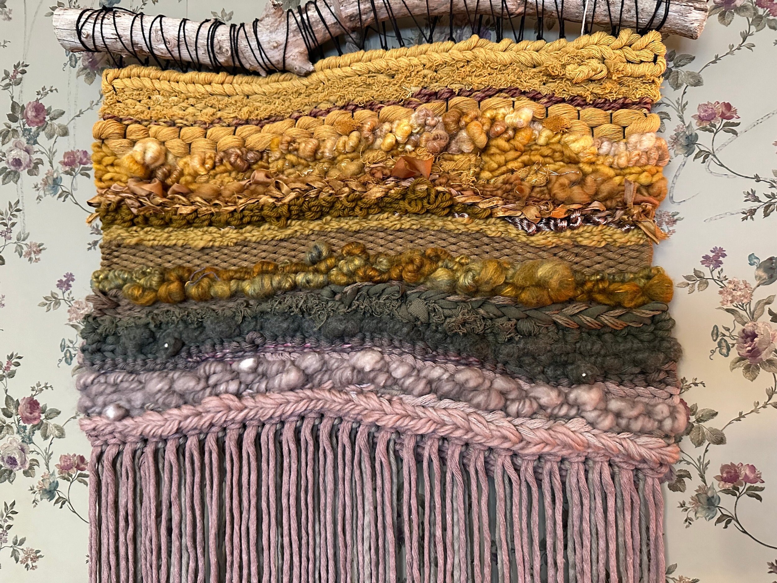 details of handmade woven tapestry in golds, greens, taupes and mauves hangs on a driftwood
