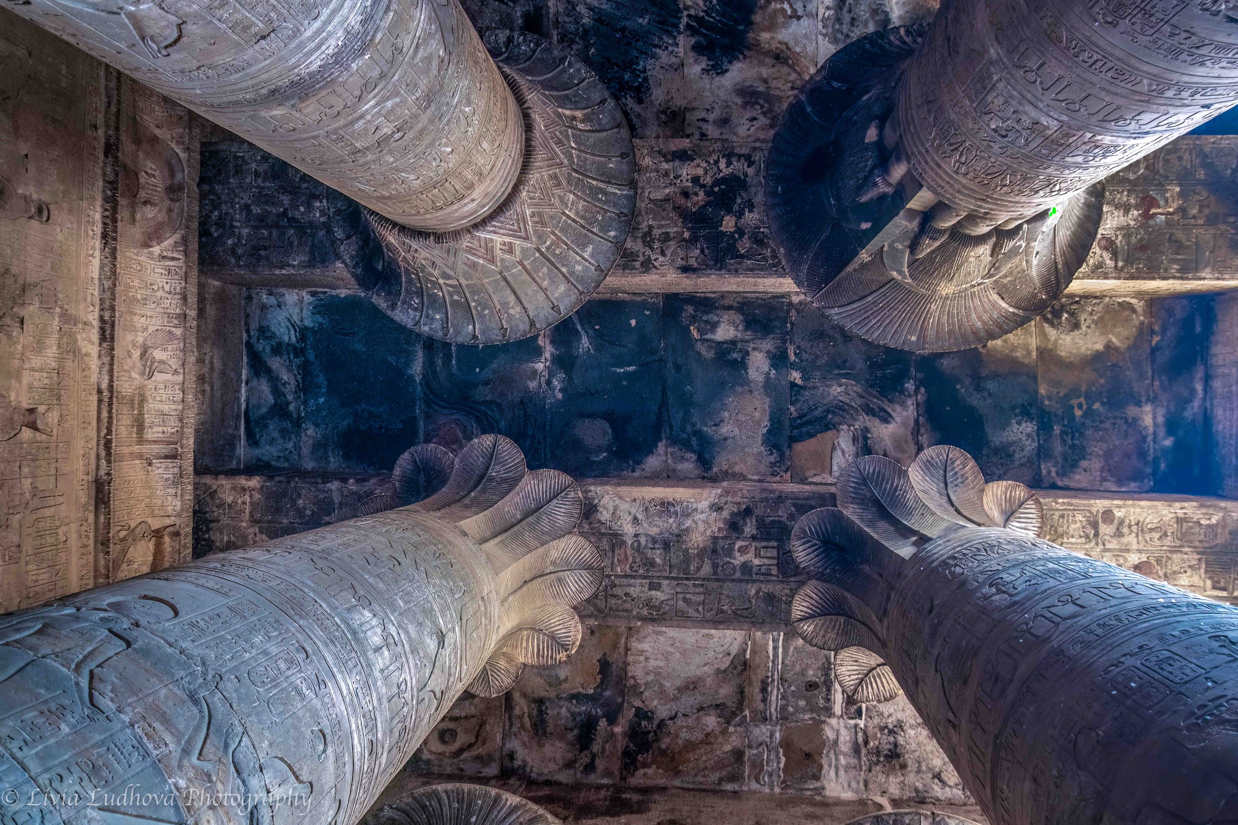 Looking up beneath the hypostyle hall at Edfu, where towering columns crowned with papyrus capitals support a ceiling darkened by centuries of incense, ritual, and time.