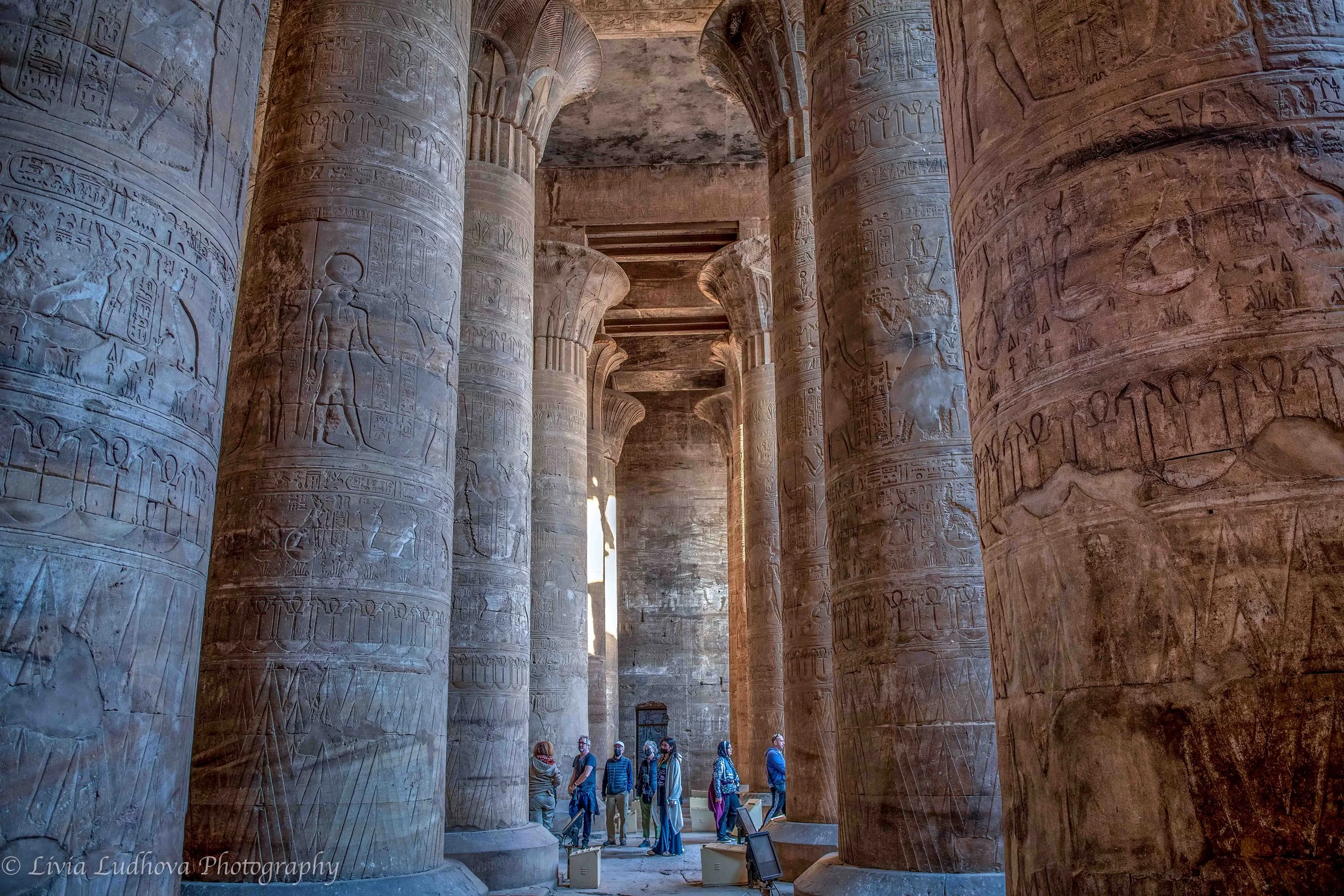 Visitors dwarfed by the massive columns of the hypostyle hall at Edfu, where towering Ptolemaic pillars once framed ritual processions leading toward the sanctuary of Horus.