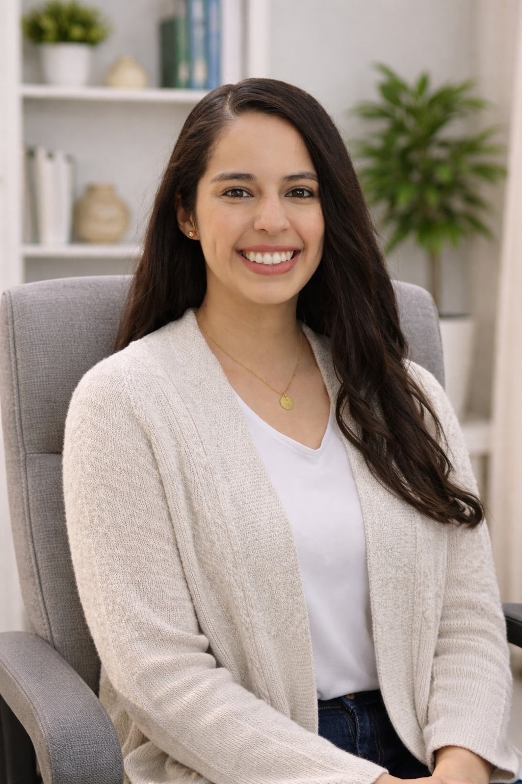 A woman with long dark hair smiling while sitting in a gray office chair in a cozy room with shelves, plants, and decorative items.