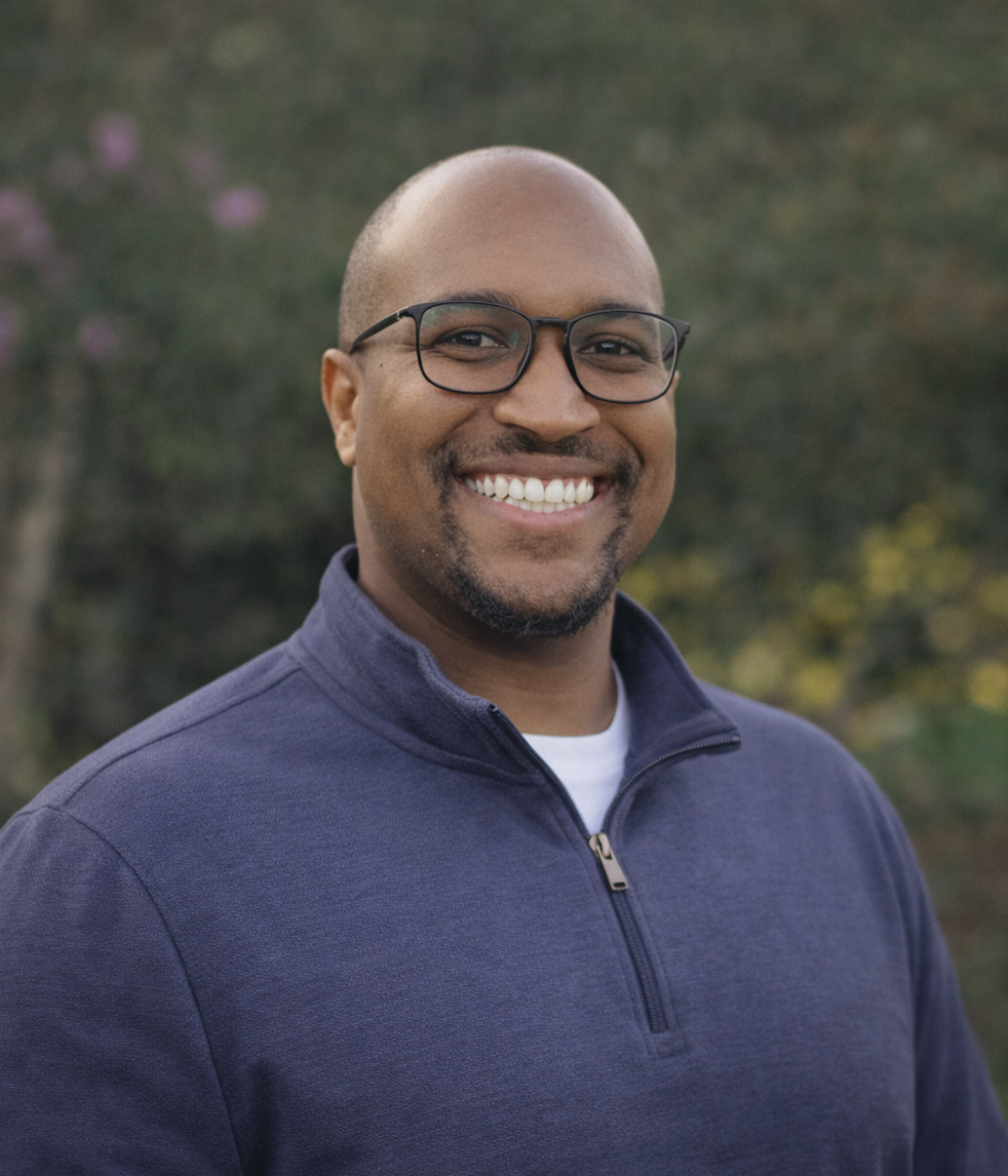 A smiling man wearing glasses and a colorful, patterned shirt, with a blurred outdoor background and people dining behind him.