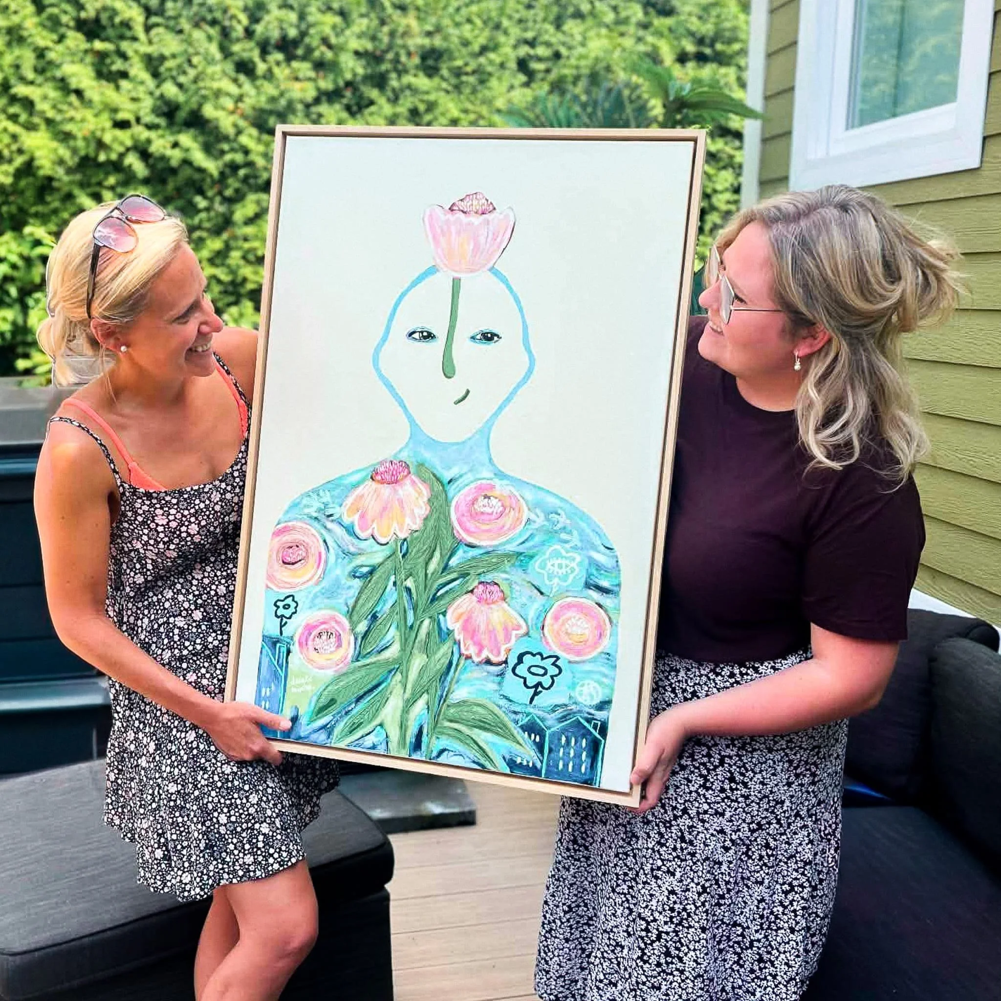 Deux femmes debout, échangeant un tableau représentant une peinture florale, en extérieur près d'une maison avec des fenêtres. La scène est lumineuse et semble être une réunion ou un événement social.