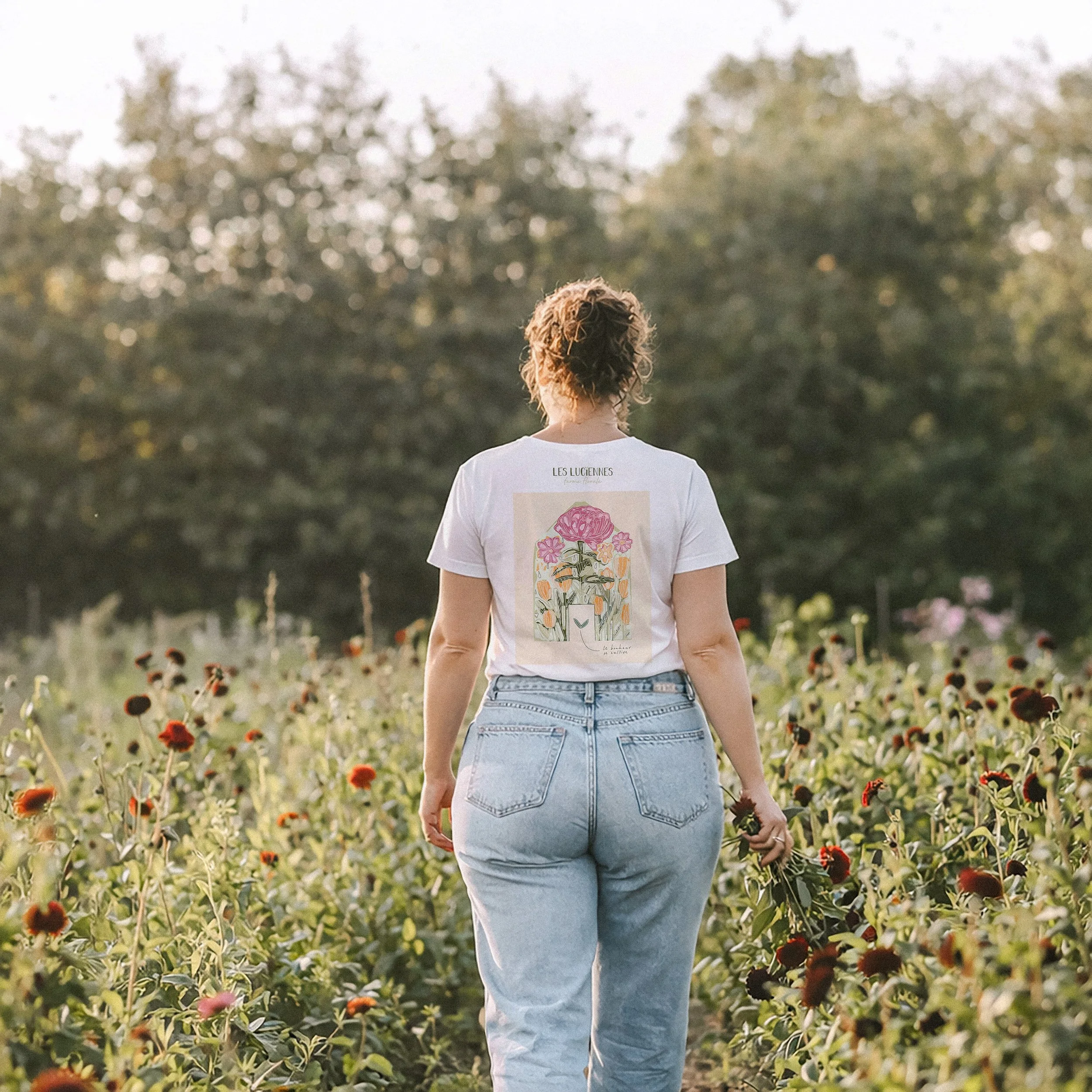 Une femme marche dans un champ de fleurs avec des arbres en arrière-plan, vue de dos.