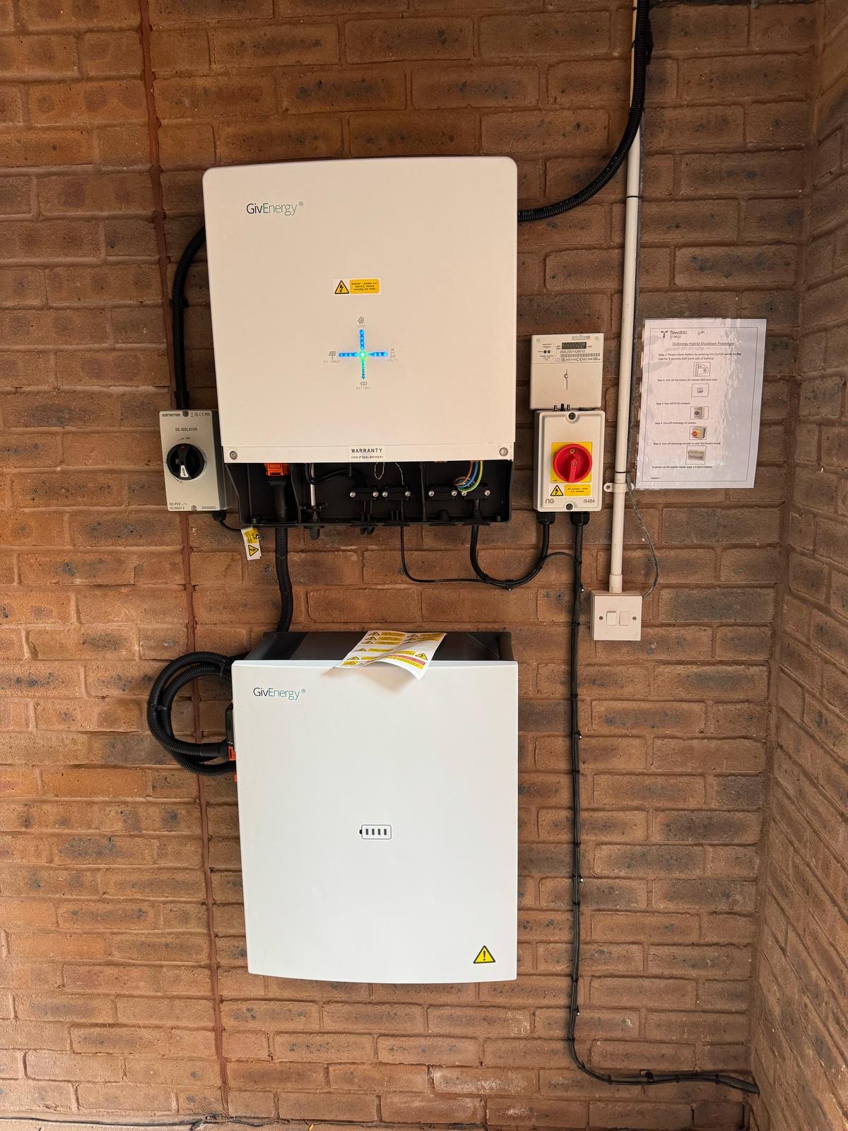 Electrical and solar power equipment mounted on a brick wall, including a white inverter, a black battery storage unit, a red emergency switch, and various cables and control panels.