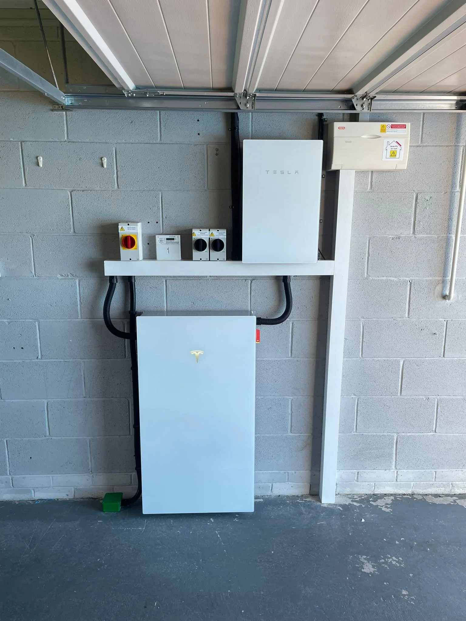 Tesla power wall energy storage system installed in a garage. It includes two white rectangular units, one mounted on a metal frame and the other lower on the wall, with various electrical disconnect switches and wiring.
