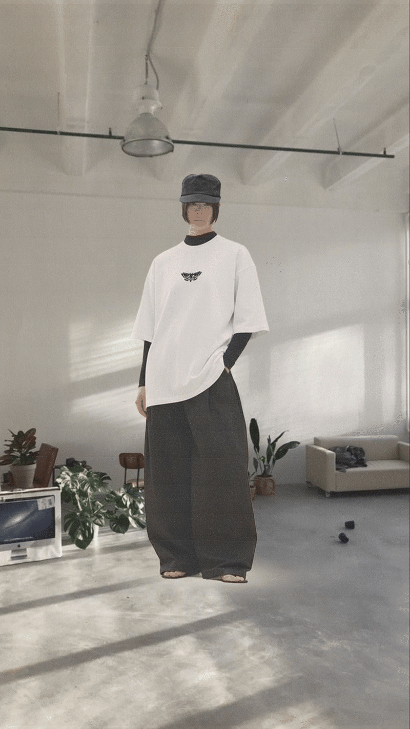 A person standing in a minimalist room with concrete floors and ceiling, wearing a black hat, a white oversized T-shirt with a small black bird logo, black long sleeves underneath, and wide-leg black pants, with plants and furniture in the background.