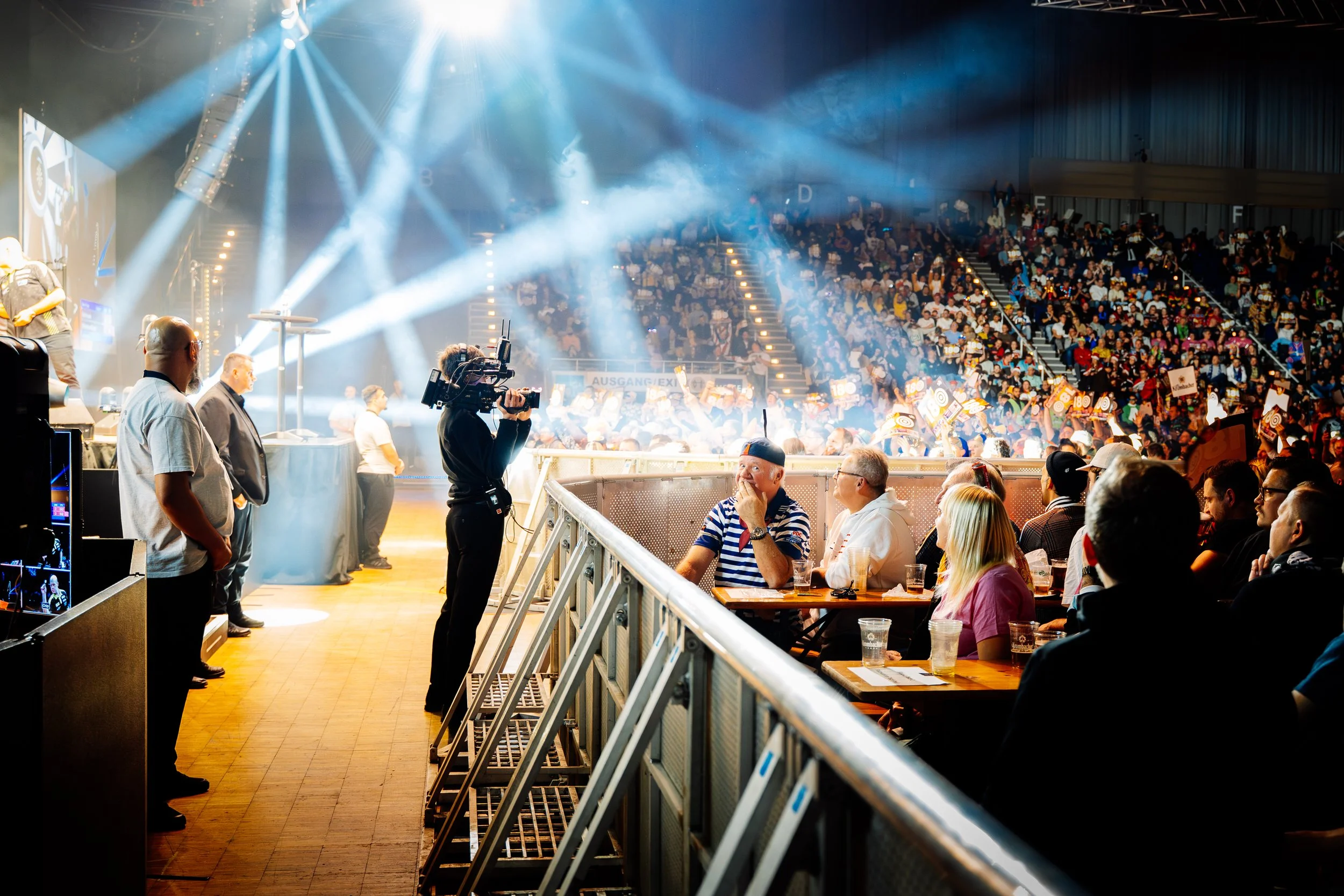 Menschen sitzen in einem Veranstaltungsraum, während auf der Bühne eine Show mit Scheinwerfern und einem Publikum im Hintergrund stattfindet.