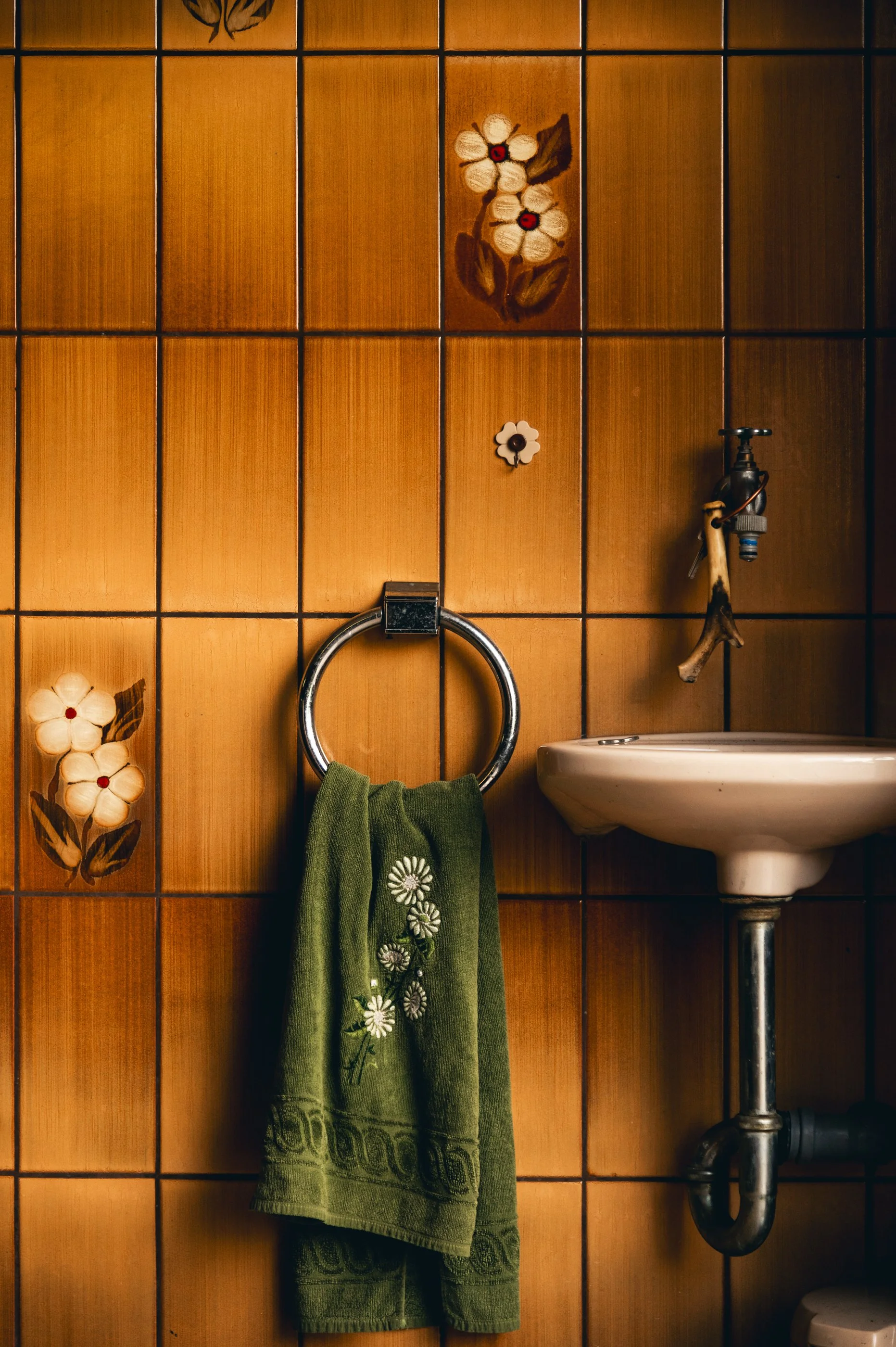 Das Bild zeigt eine kleine, vintageartige Ecke eines Badezimmers mit braunen, blumenteierten Fliesen an der Wand, einem grünen Handtuch, einem kleinen Waschbecken und einem Wasserhahn.
