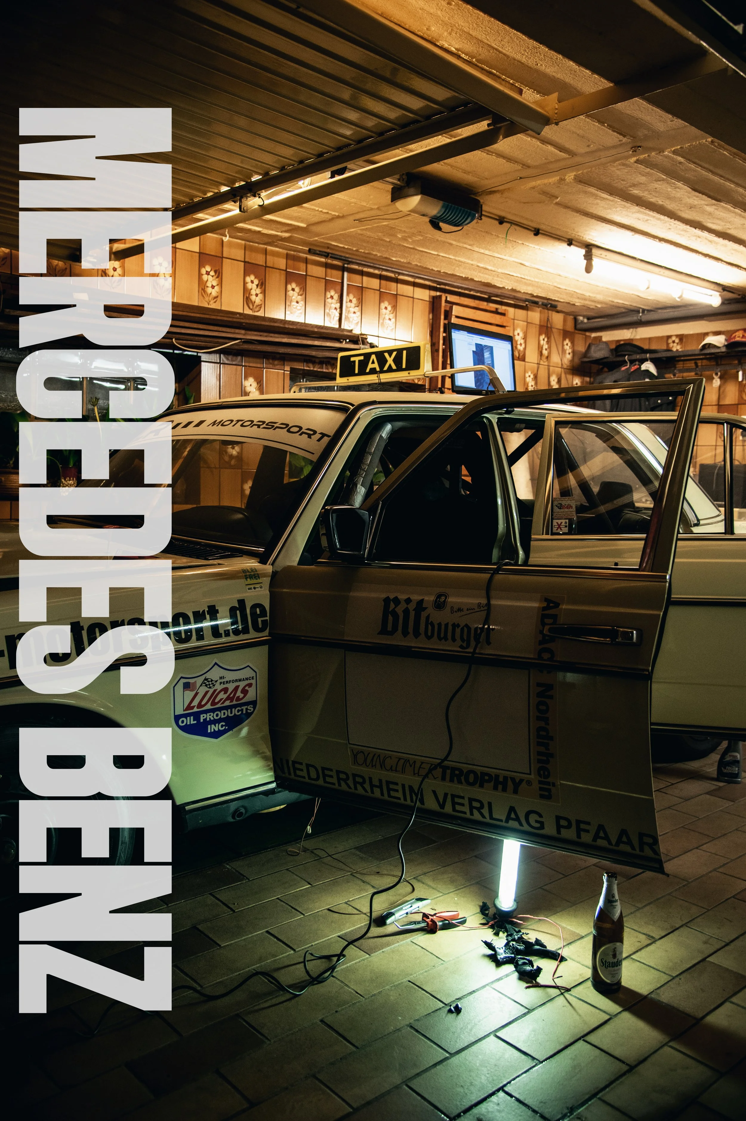 Ein Oldtimer-Taxi in einer Werkstatt, umgeben von Werkzeugen, einem Laptop und einer Getränkeflasche, mit großen weißen Text über dem Bild, der sagt "LUEBSTENZ".
