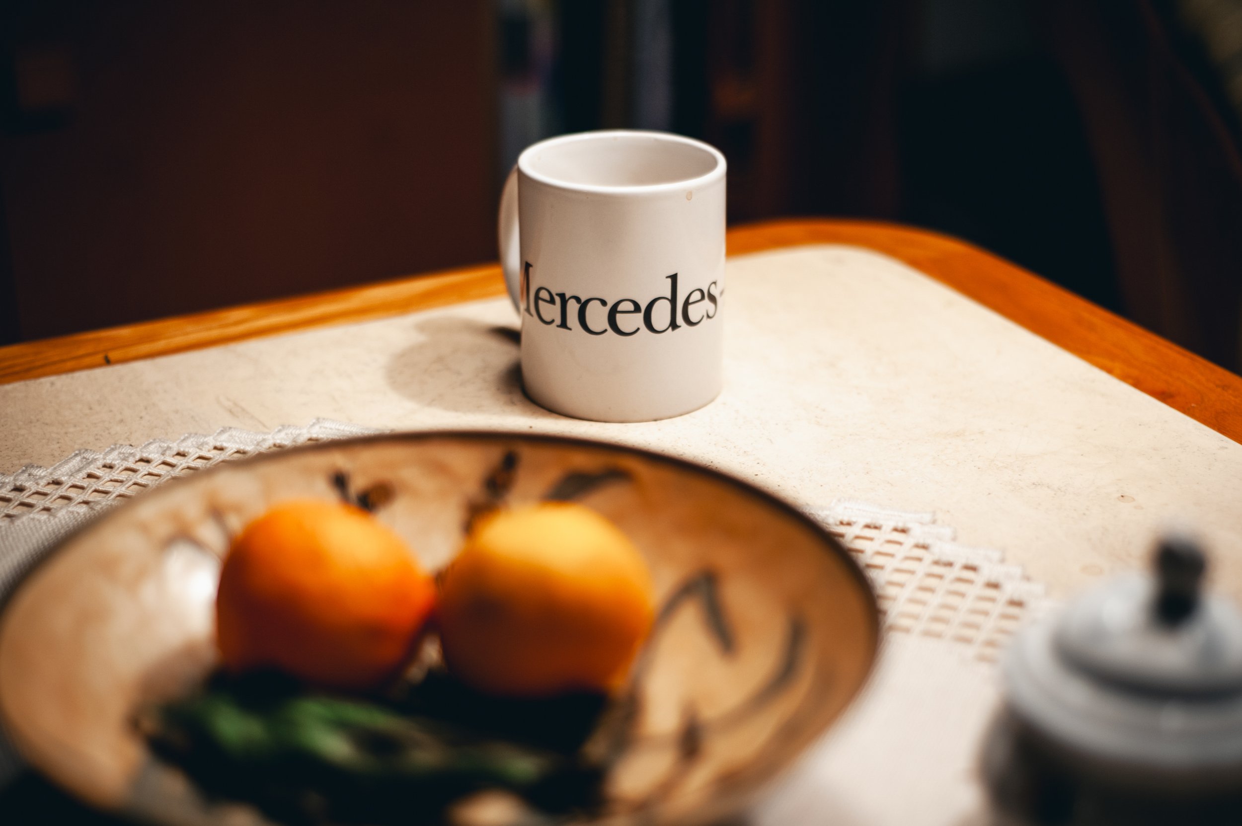 Weiße Tasse mit Mercedes-Benz Logo auf einem Holztisch, im Vordergrund eine Schale mit zwei Äpfeln und grünem Blatt, im Hintergrund eine kleine Dose mit Deckel.
