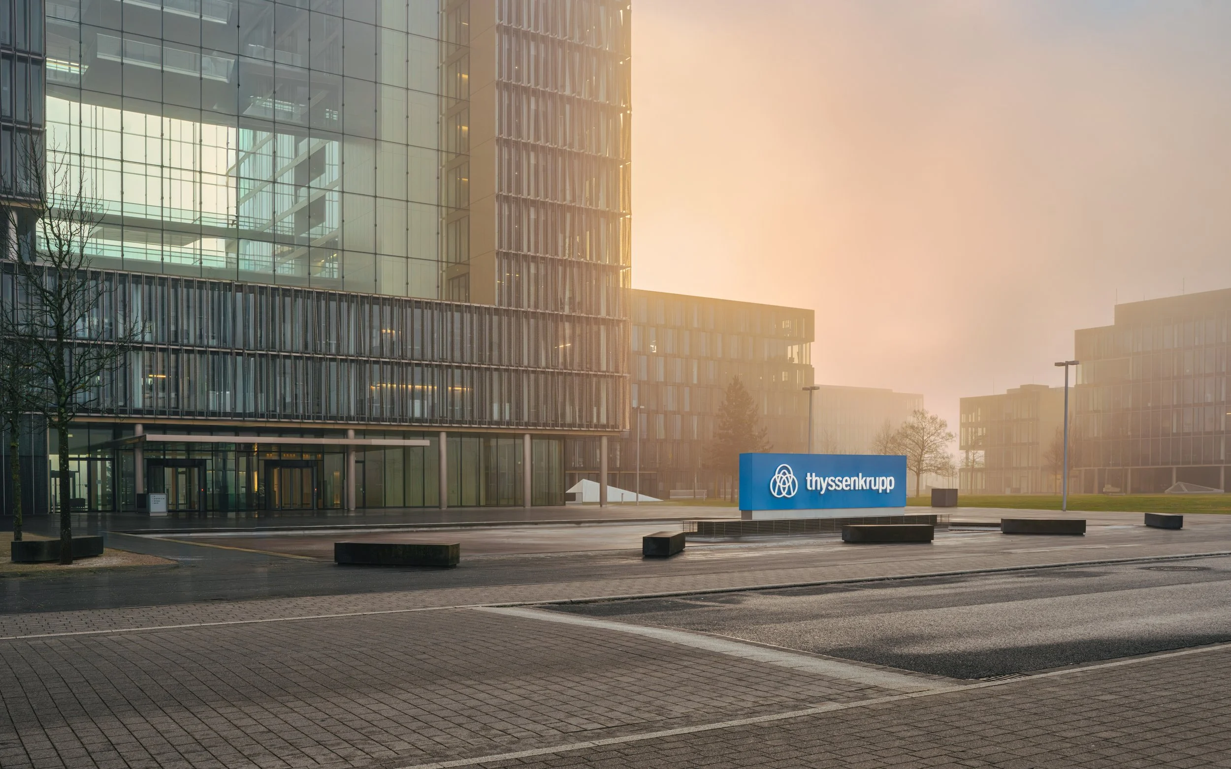 Modernes Firmengebäude mit einem blauen Schild, das den Schriftzug 'thyssenkrupp' trägt, bei Sonnenaufgang oder -untergang in einer ruhigen, urbanen Umgebung.