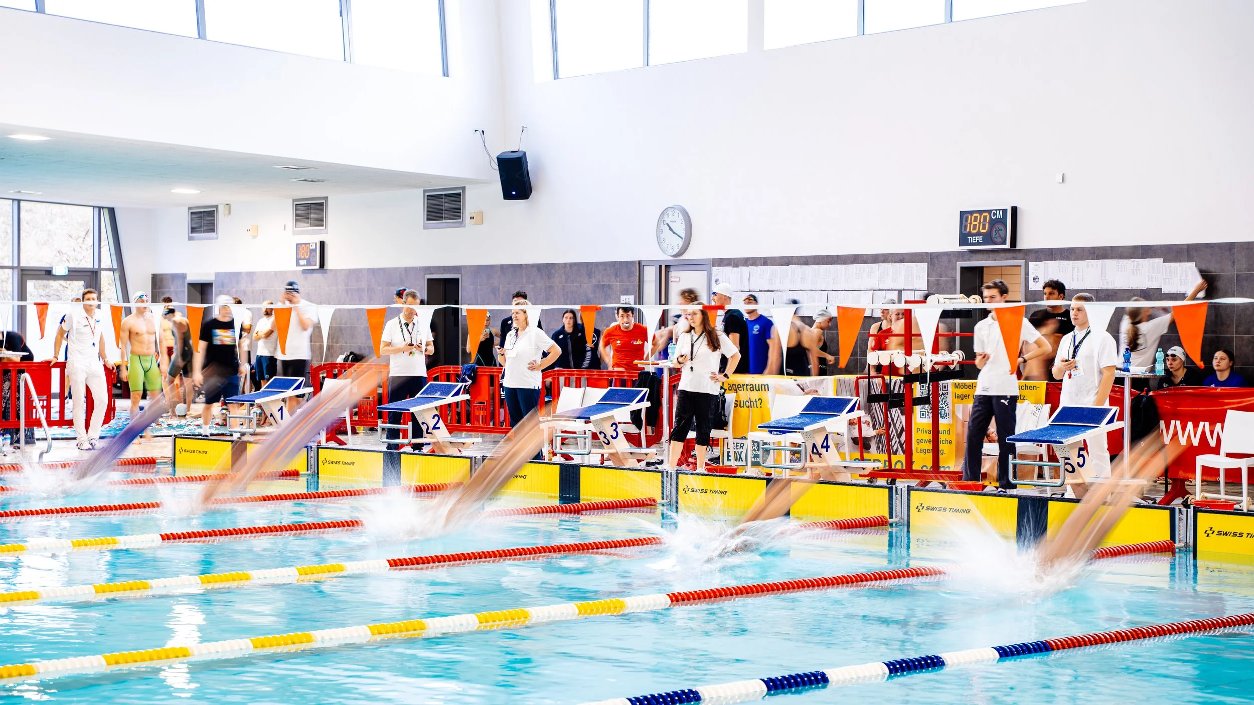 Schwimmer starten im Schwimmbecken bei einem Wettkampf, während Trainer und Betreuer am Beckenrand stehen und zuschauen.