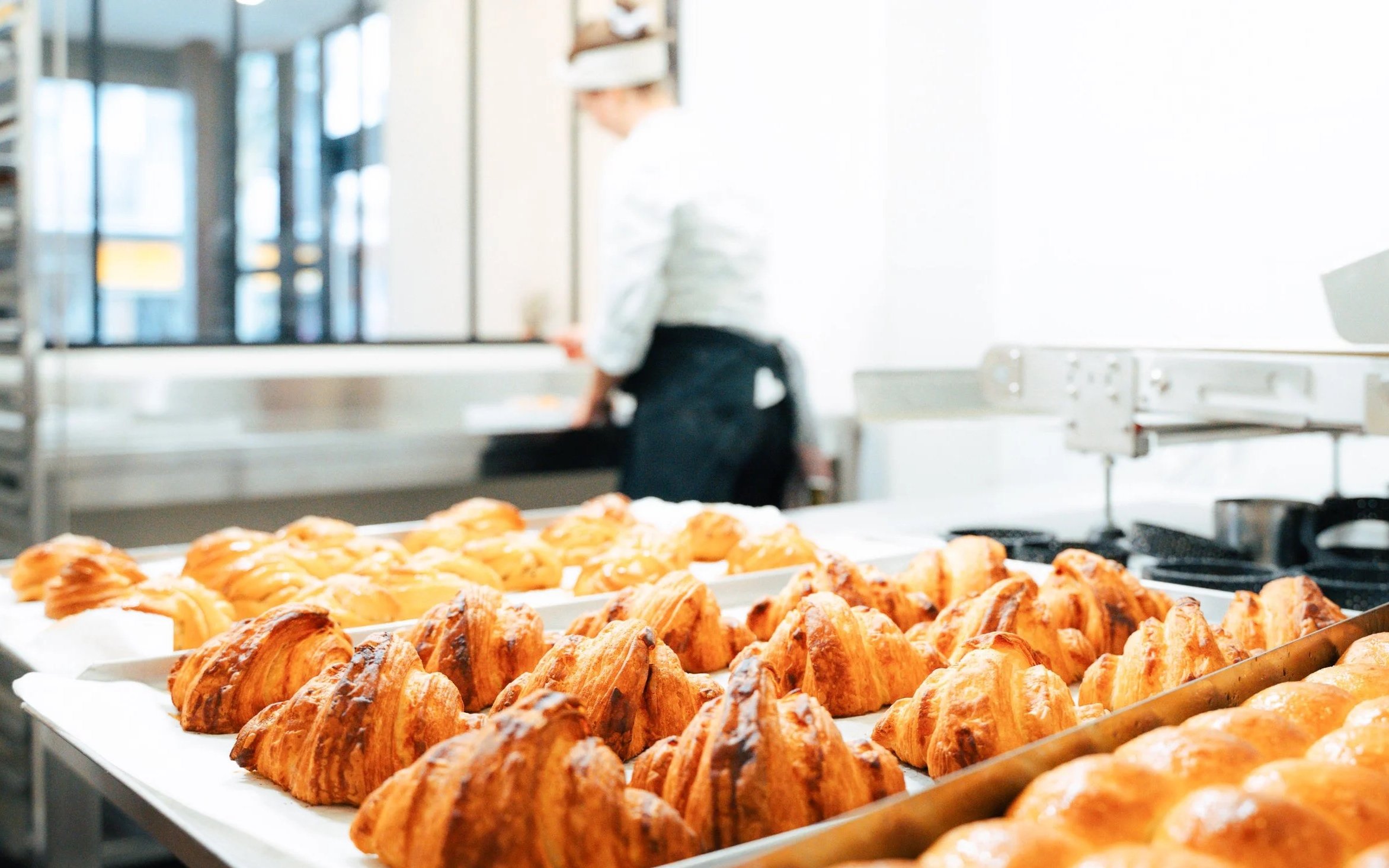 Frisch gebackene Croissants auf einem Blech in einer Bäckerei, mit einer Bäckerin im Hintergrund.