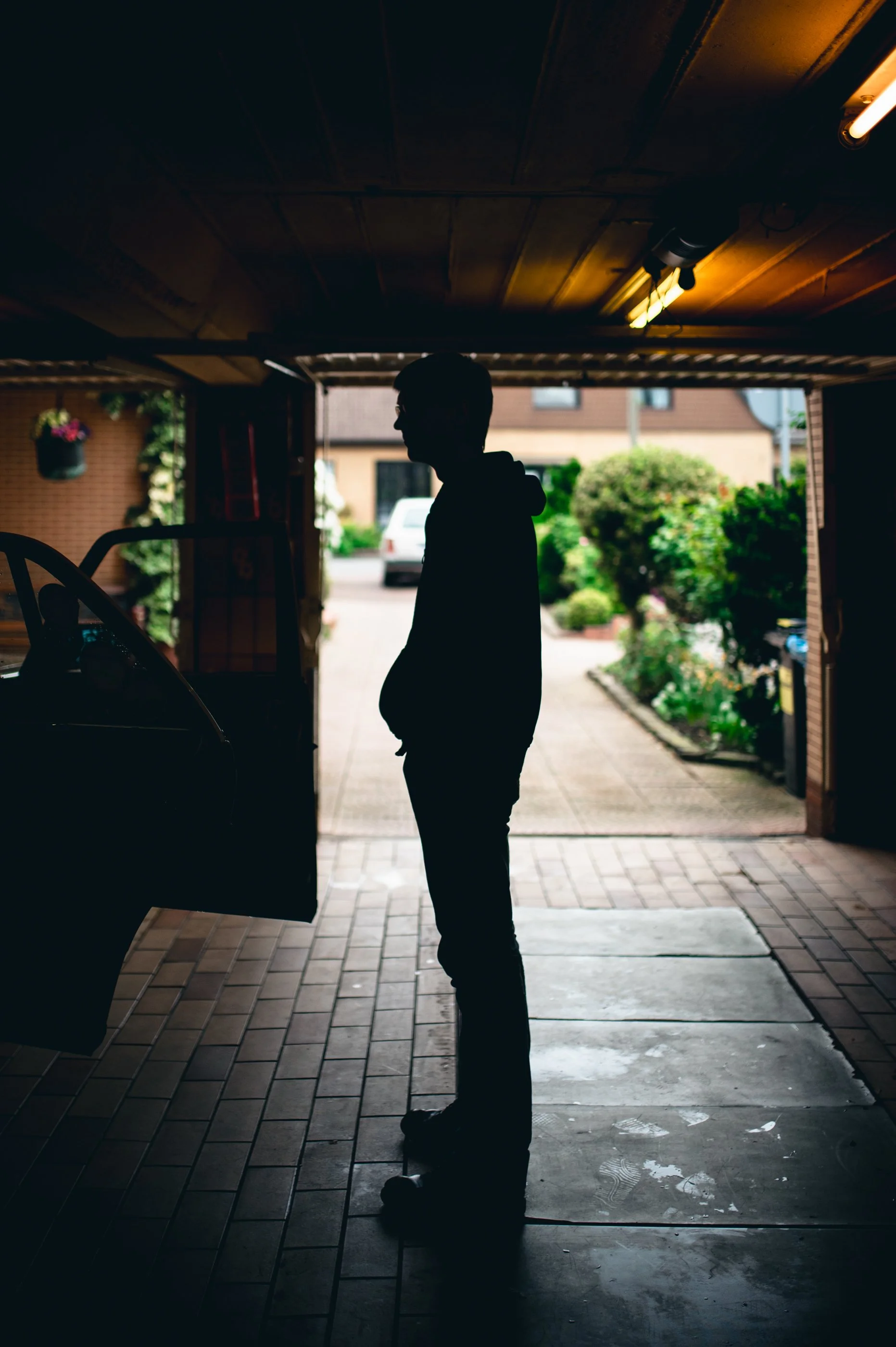 Silhouette eines Mannes steht in einer Garage vor der Ausfahrt, im Hintergrund zeigt sich ein Garten und ein Haus.