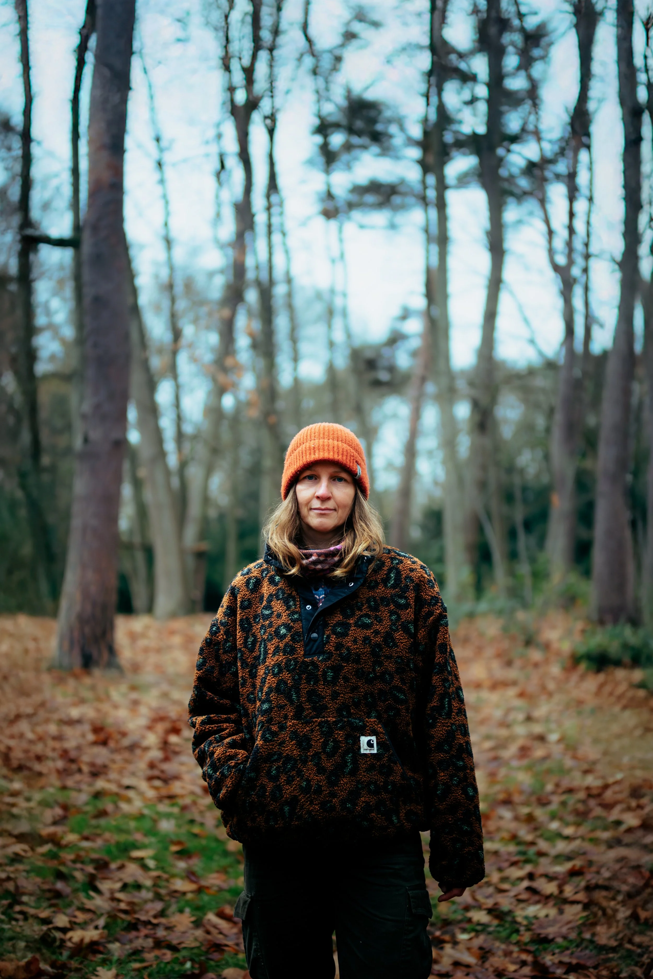Eine Frau steht in einem herbstlichen Wald mit Laub auf dem Boden. Sie trägt eine orange Mütze, eine braun-schwarz gefleckte Jacke und schwarze Hosen.