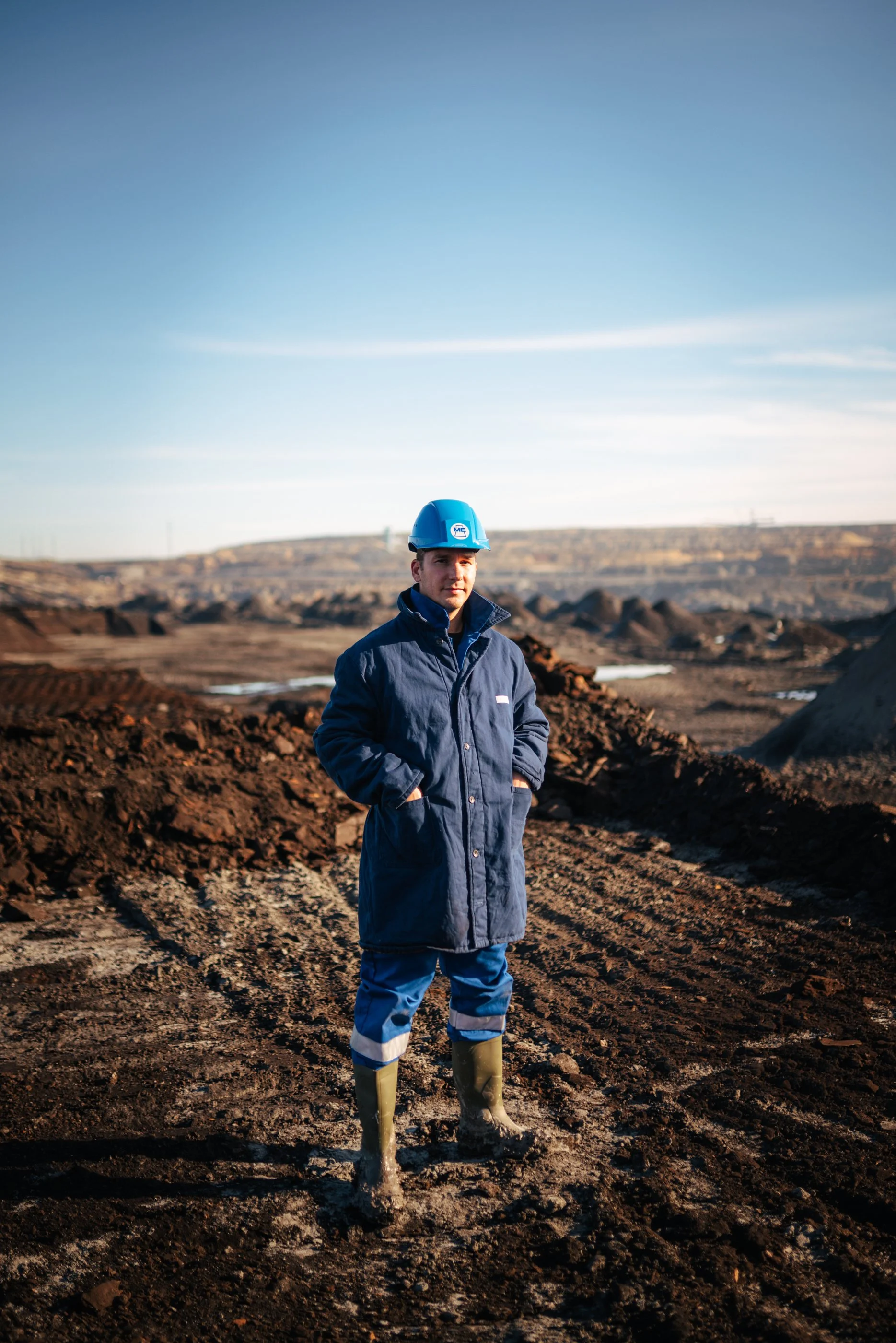 Ein Mann steht auf einer Baustelle im Freien, trägt einen blauen Schutzhelm, eine dunkle Jacke und Gummistiefel vor einem Hintergrund von Erdausgrabungen und weitem Himmel.