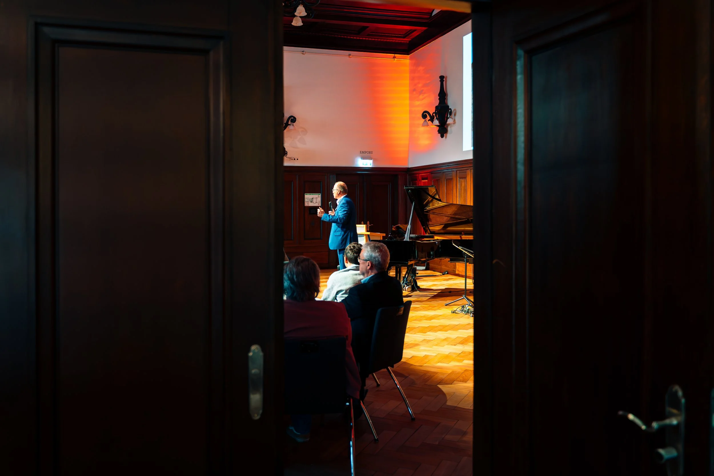 Ein Konzertsaal mit einem stehenden Mann in blauen Anzügen, der auf einer Bühne spricht, während mehrere sitzende Zuhörer zuhören. Es gibt ein Klavier auf der Bühne und der Raum ist mit Holzvertäfelungen ausgekleidet.