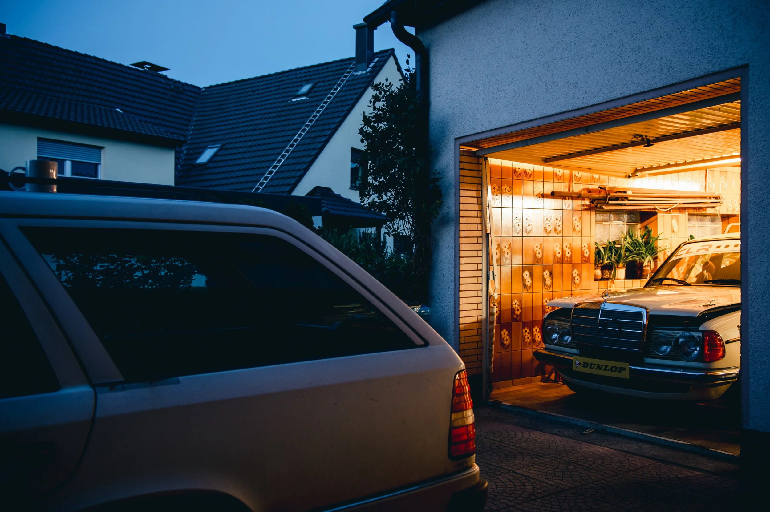 Ein geöffneter garagenähnlicher Raum, in dem ein alter Mercedes-Benz Auto steht, umgeben von Pflanzen und mit einer warmen Beleuchtung. Im Vordergrund ist ein Teil eines anderen Autos sichtbar.