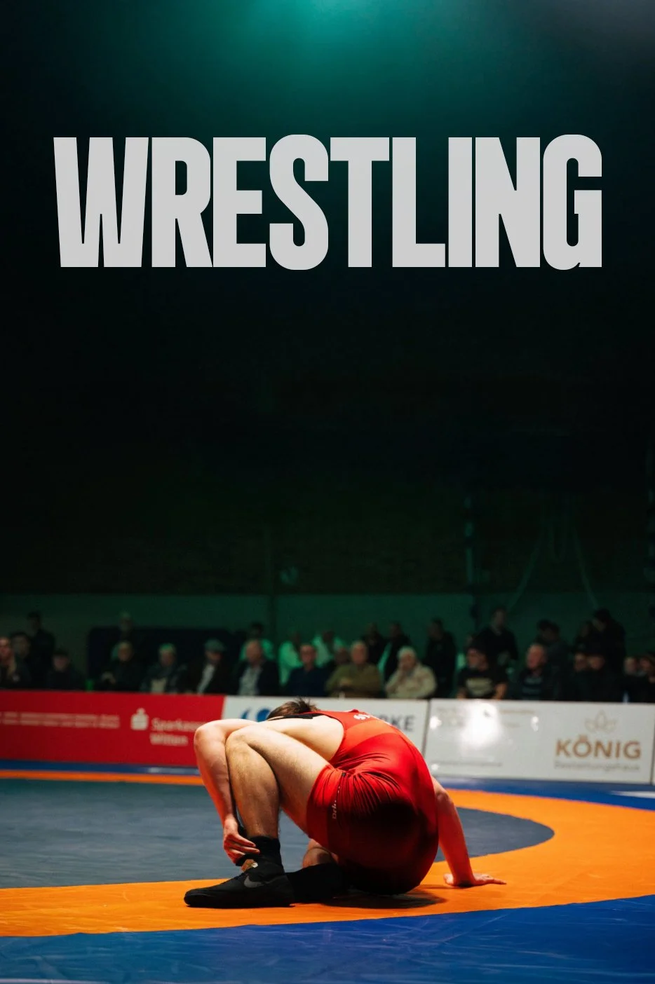 Ein Ringer in einer roten Uniform in einer Wrestling-Arena, umgeben von Zuschauern, die im Hintergrund sitzen.