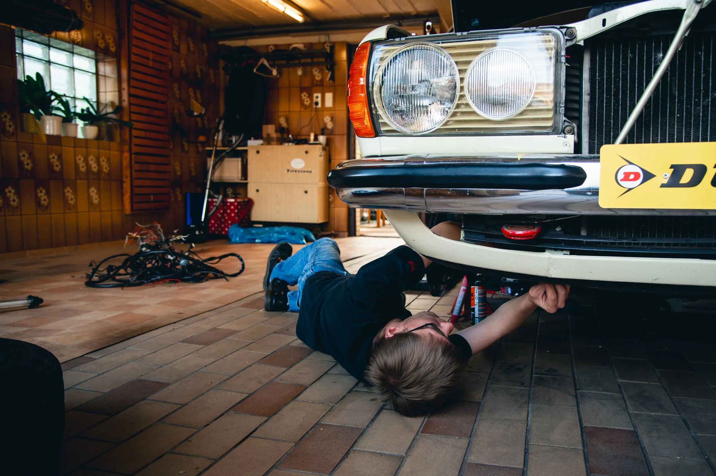 Eine Person repariert ein Auto in einer Garage, liegt unter dem Fahrzeug und arbeitet an der Unterseite. Im Hintergrund sind Werkzeuge und Kabel auf dem Boden verteilt.