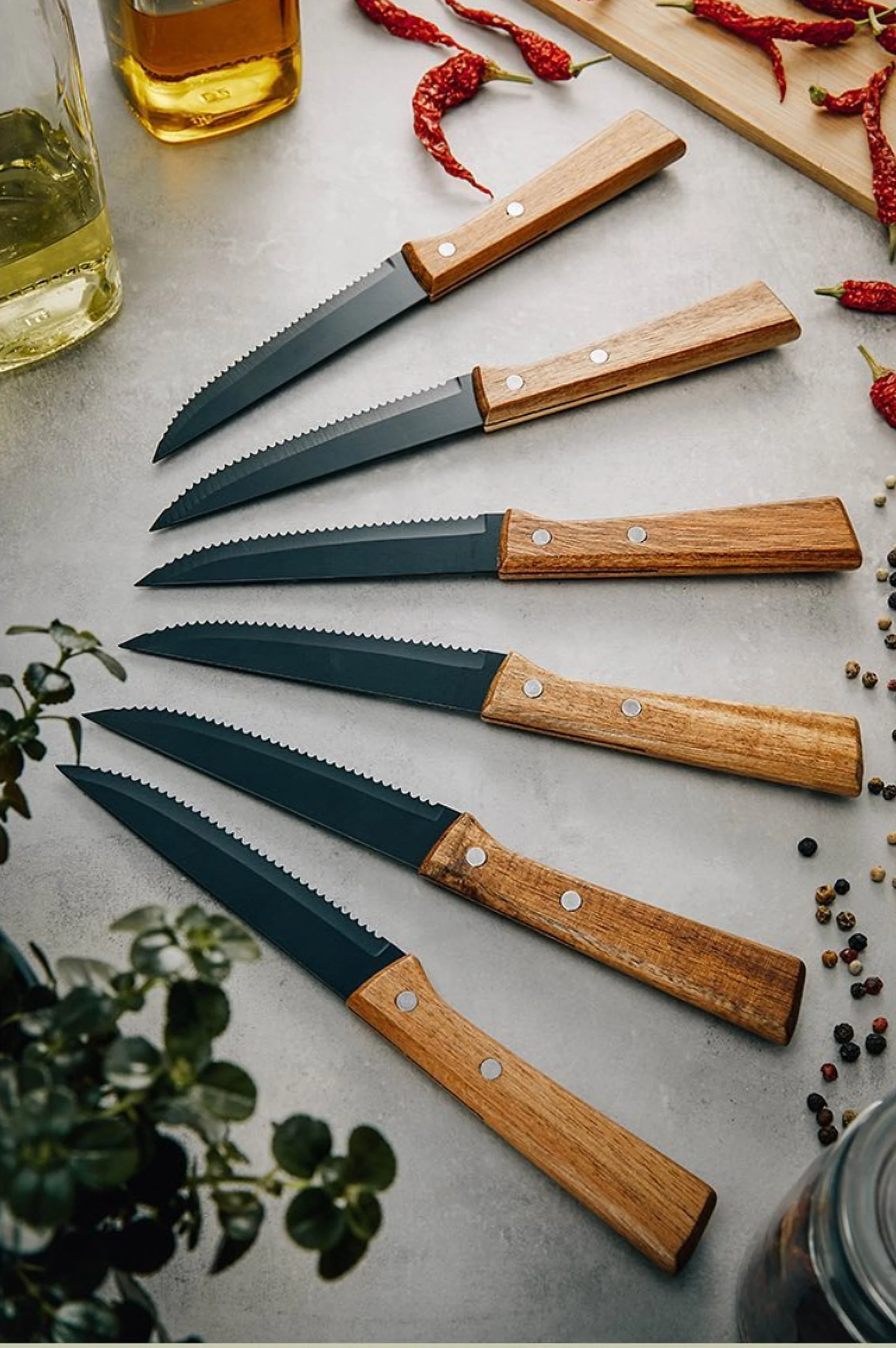 Sechs Kochmesser mit schwarzen Klingen und Holzgol stellen auf einer Küchenarbeitsplatte nebeneinander, umgeben von Gewürzen, Chilis, Ölen, Pflanzen und einem Glas.