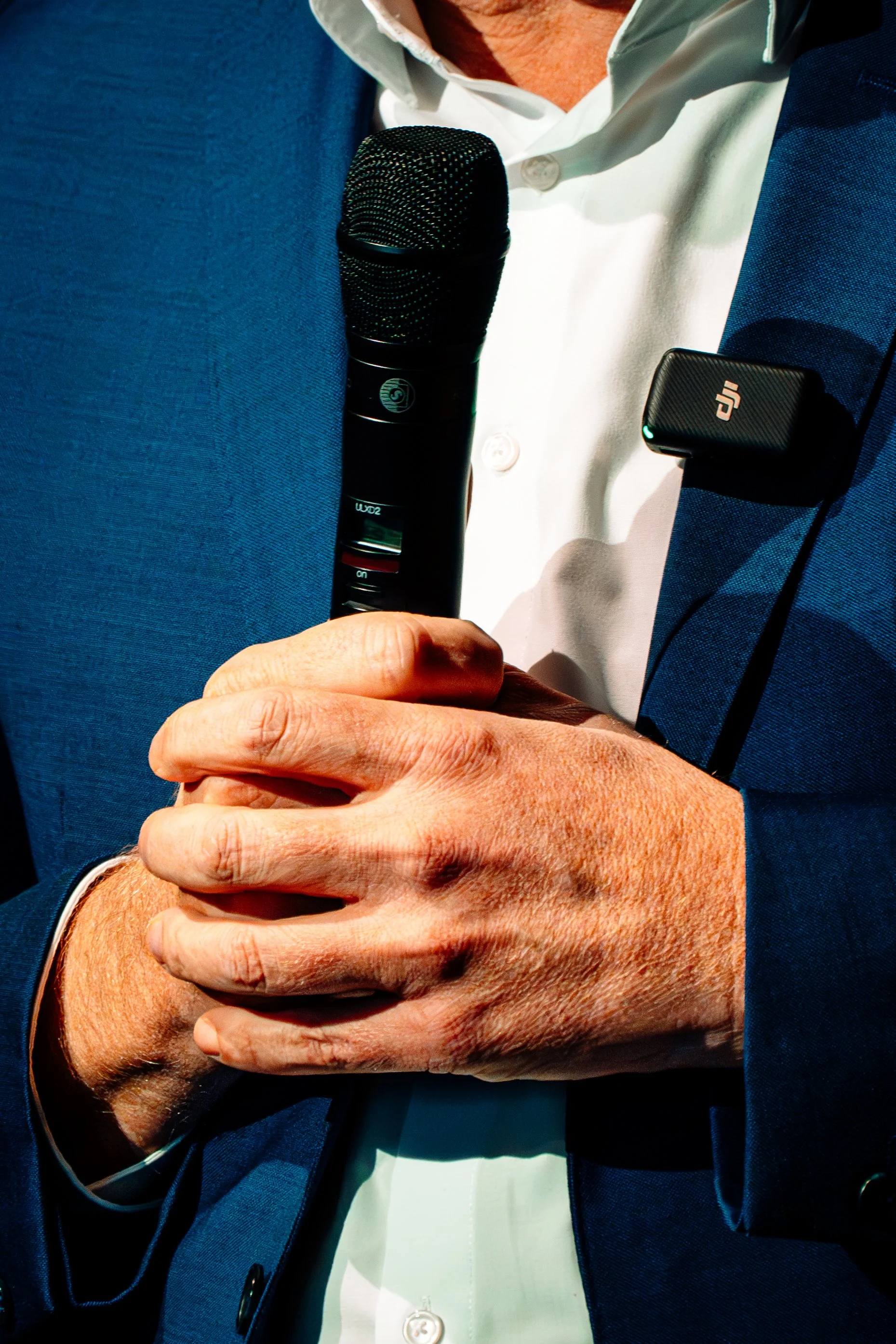 Nahaufnahme eines Mannes in Anzug, der ein Mikrofon in der Hand hält, mit einem DJI-Mikrofon und einem Funkmikrofon befestigt an seinem Anzug.