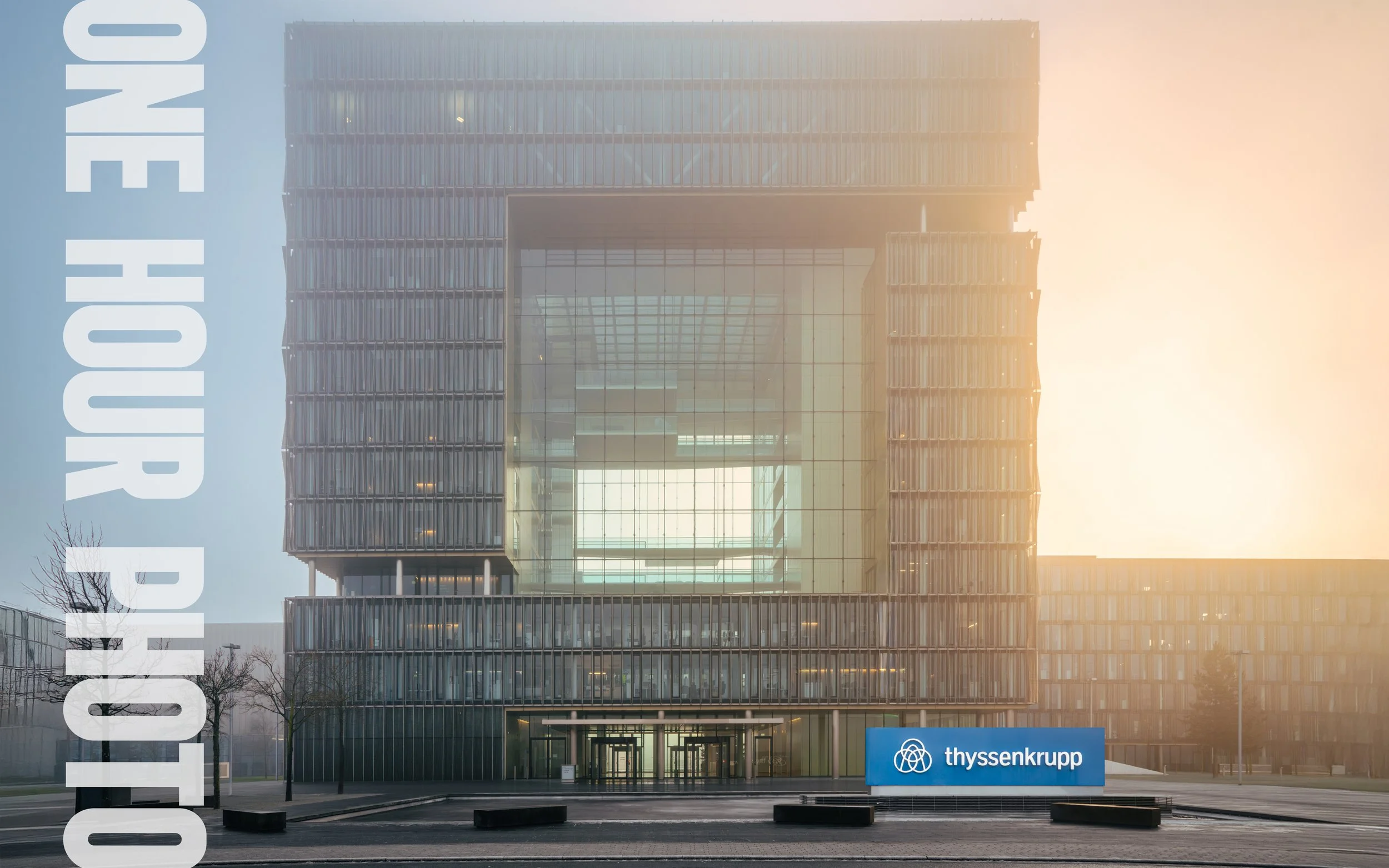 Das Bild zeigt das moderne Bürogebäude der Thyssenkrupp-Unternehmenszentrale bei Sonnenaufgang oder Sonnenuntergang, mit einem großen blauen Schild mit dem Thyssenkrupp-Logo im Vordergrund.